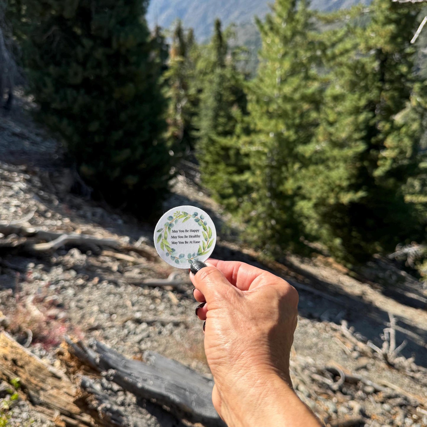 Hand holding a 2" round blue sticker with Buddhist mettā prayer text, may you be happy may you be healthy may you be at ease, with nature-themed design against a forest background.