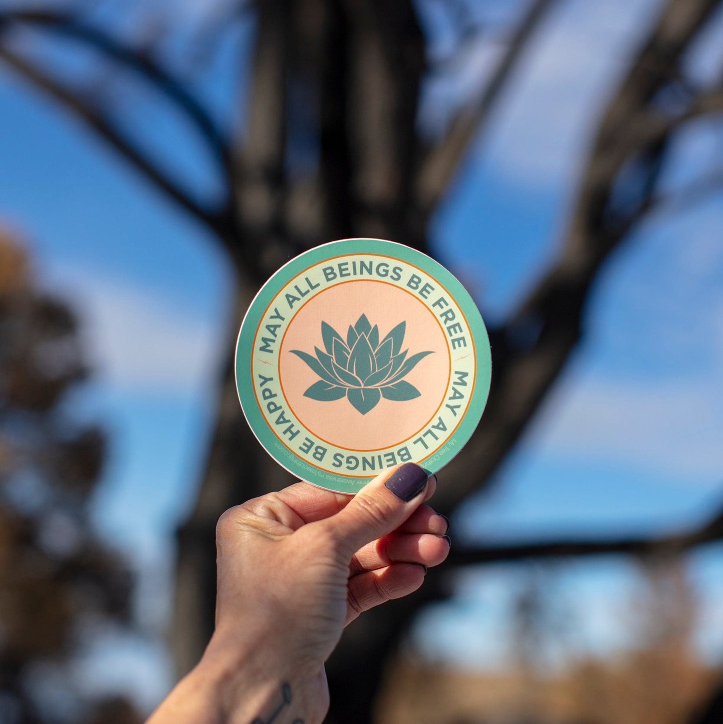 Hand holding a 4" circular sticker with a lotus flower design and text that reads, may all beings be free may all beings be happy, against a blurred natural background.