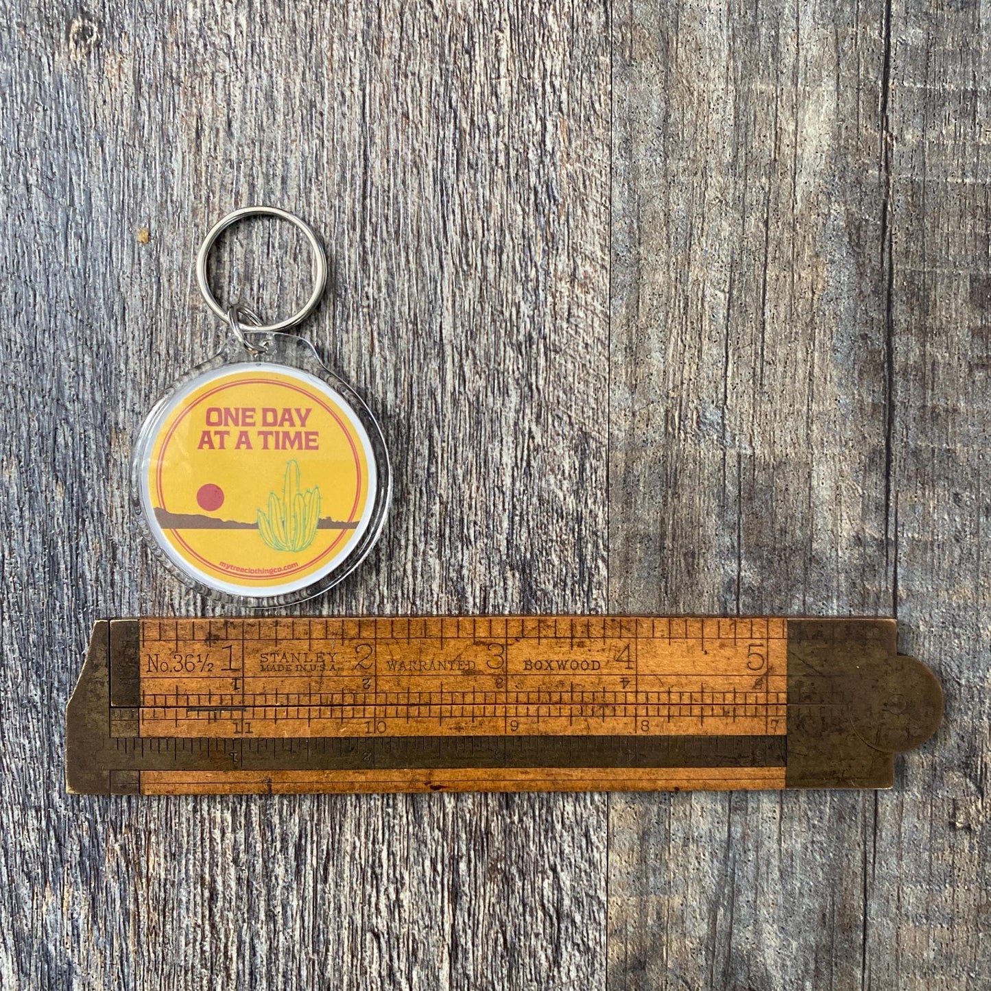 Keychain with 'One Day at a Time' text on a wooden ruler against a wooden background.