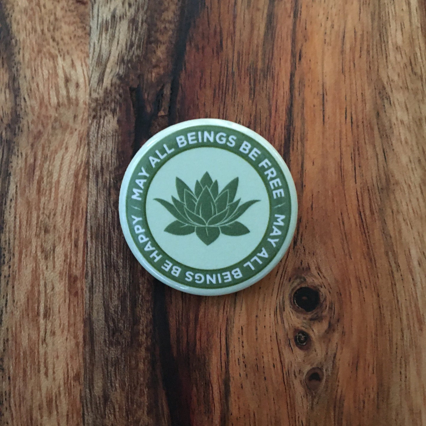 Light green 1.25" circular Buddhist pin back button with text, may all beings be free may all beings be happy, with a lotus symbol design and text on a wooden surface.