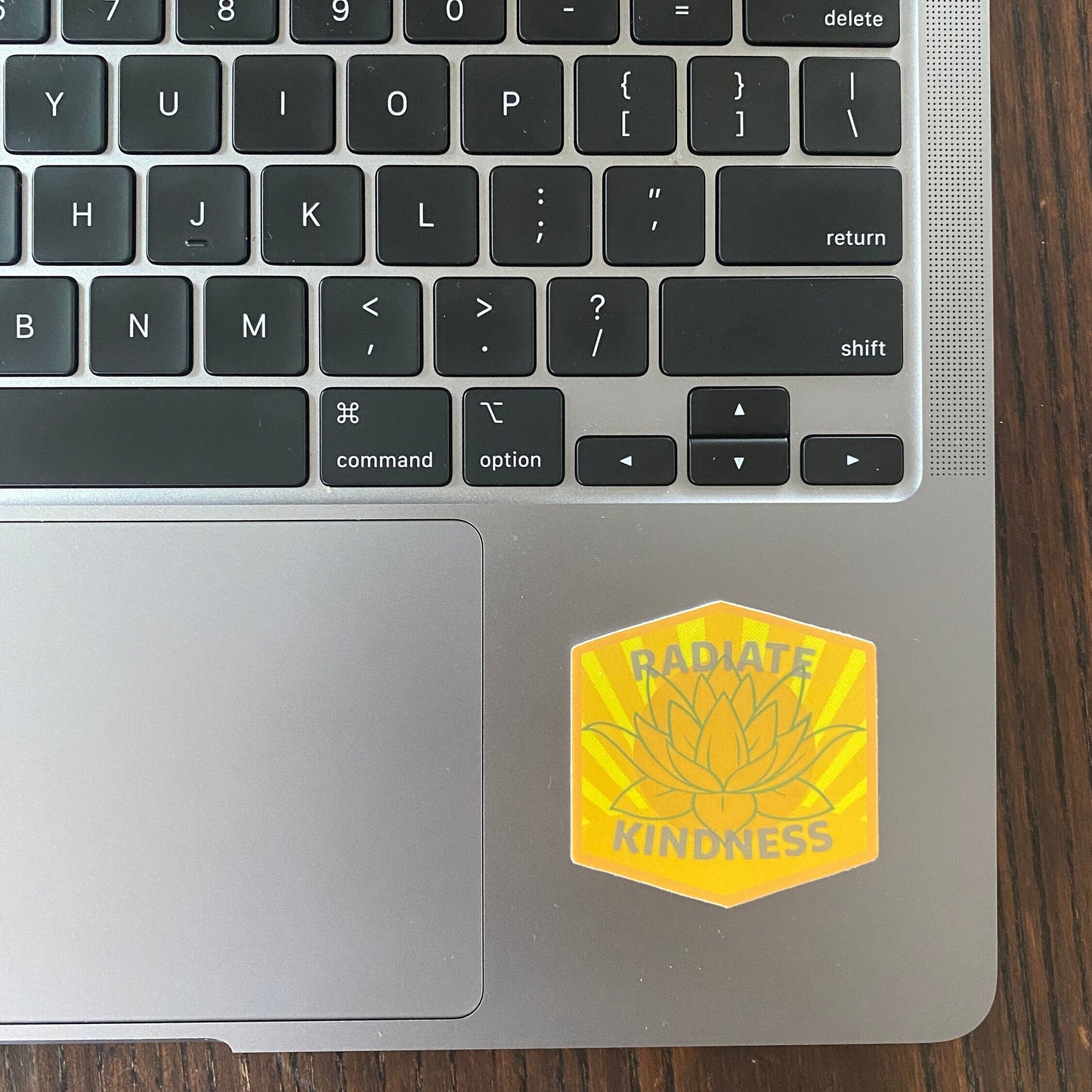 Laptop keyboard with a 'Radiate Kindness' sticker on a wooden surface