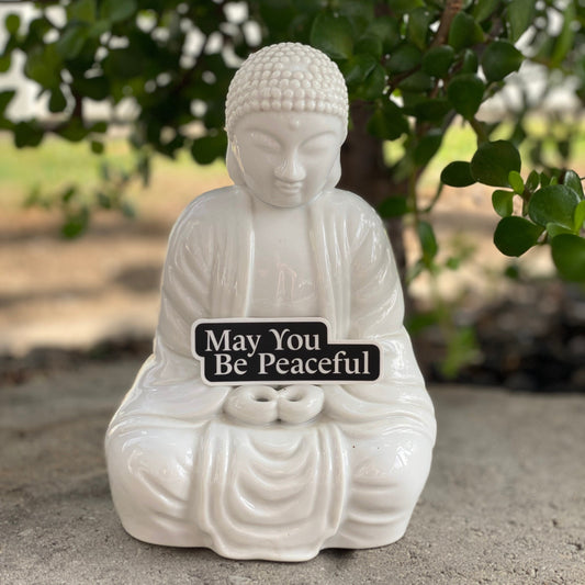 White Buddha statue with a black and white metta inspired Buddhist mantra sticker that reads 'May You Be Peaceful.'