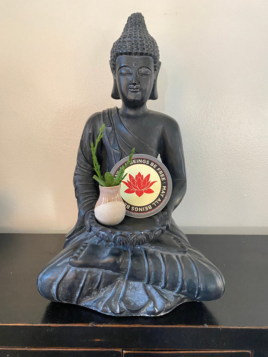 Buddha statue with a small plant and a circular sticker with a red lotus flower design and text may all beings be free may all beings be happy, on a black surface.