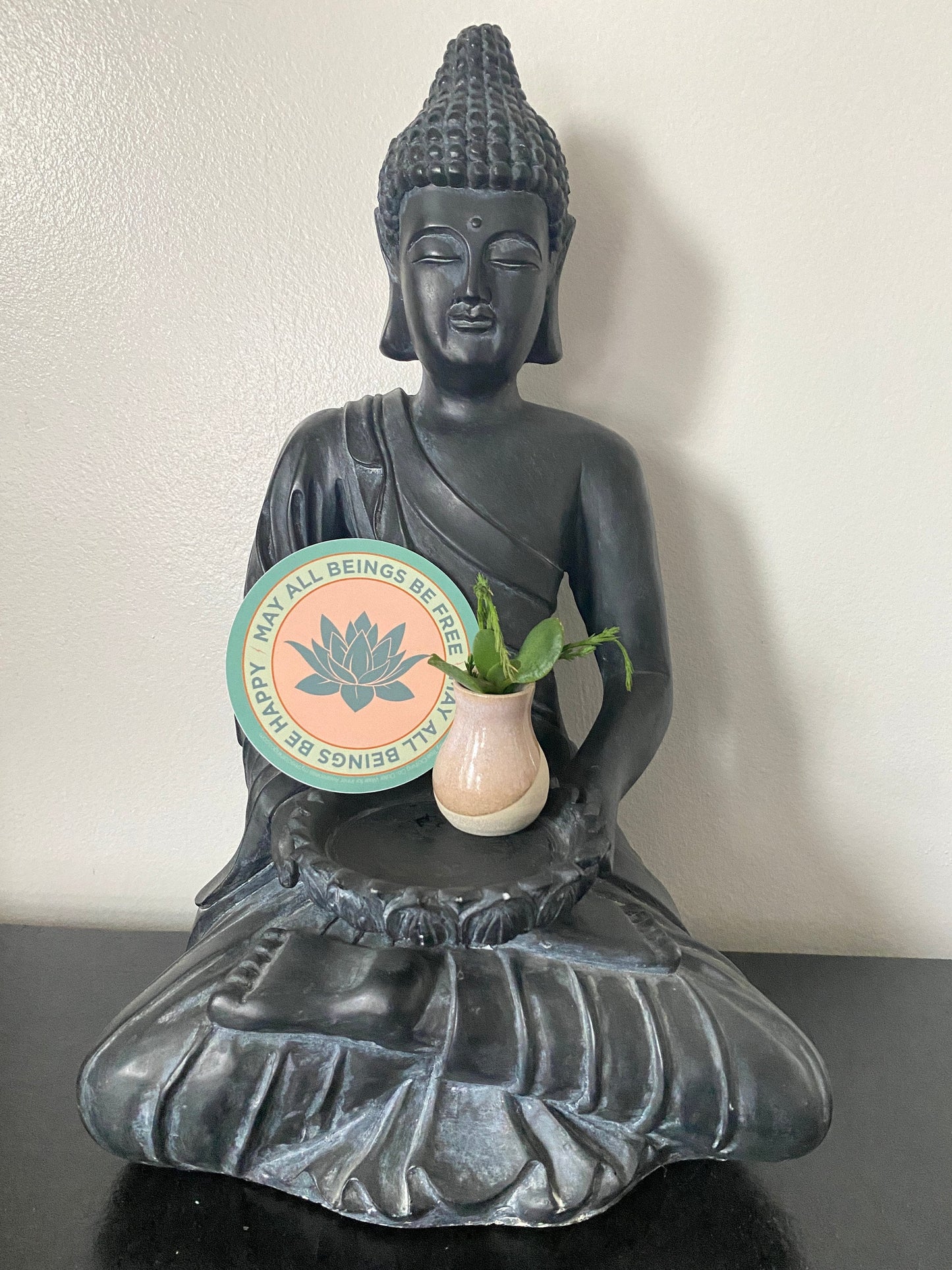 Black Buddha statue holding a small plant with a circular sticker with a lotus flower design and text that reads, may all beings be free may all beings be happy.