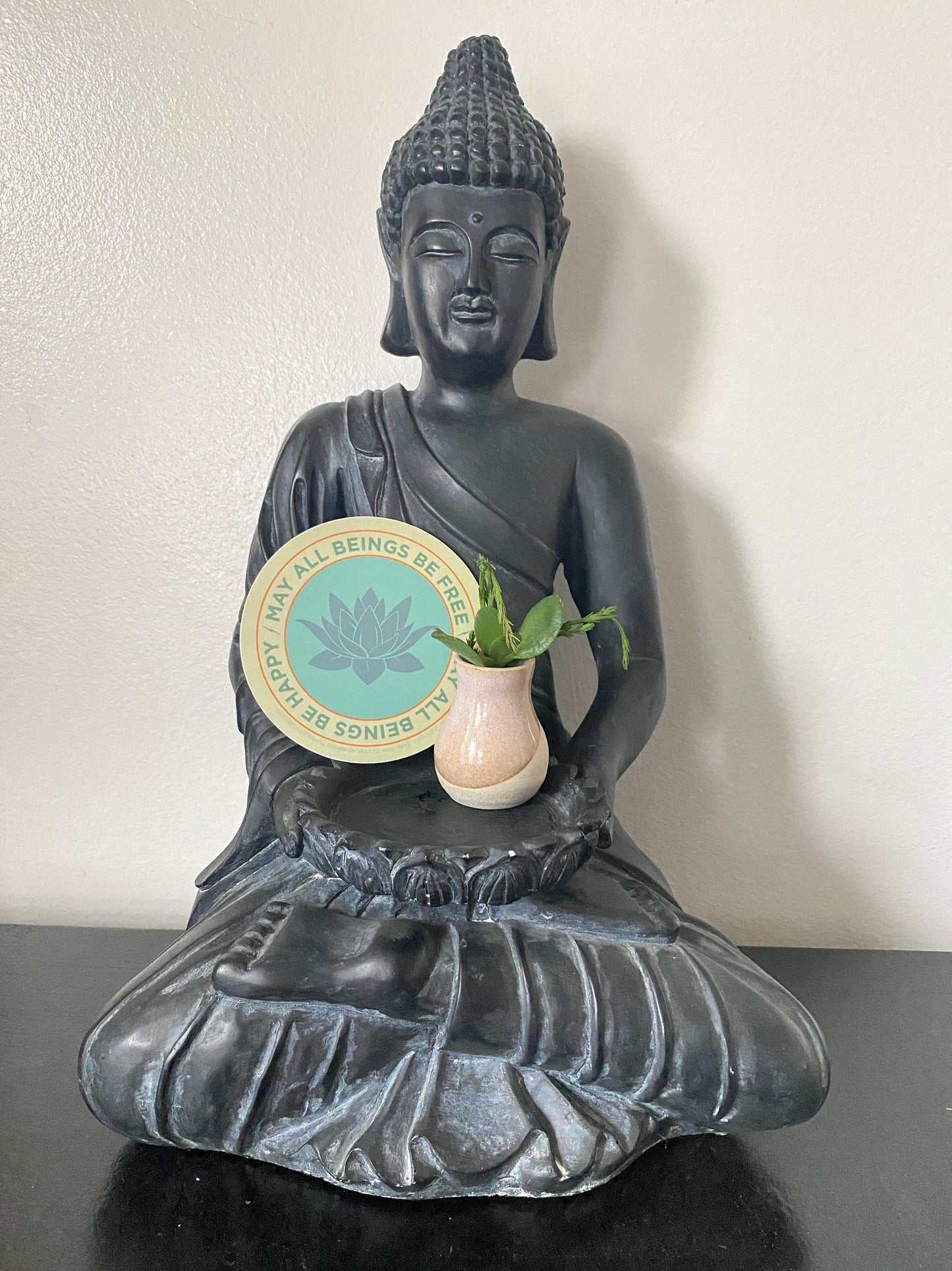 Black Buddha statue holding a vase with a plant on a white background and a 4" round lotus sticker that reads, may all beings be free may all beings be happy.