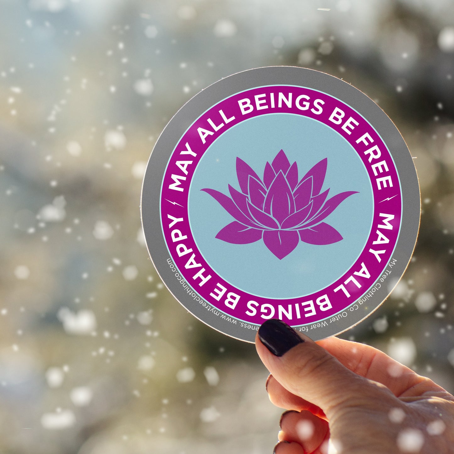 4" circular Buddhist sticker with a purple lotus flower and text, may all beings be free may all beings be happy, held by a hand against a blurred natural background.