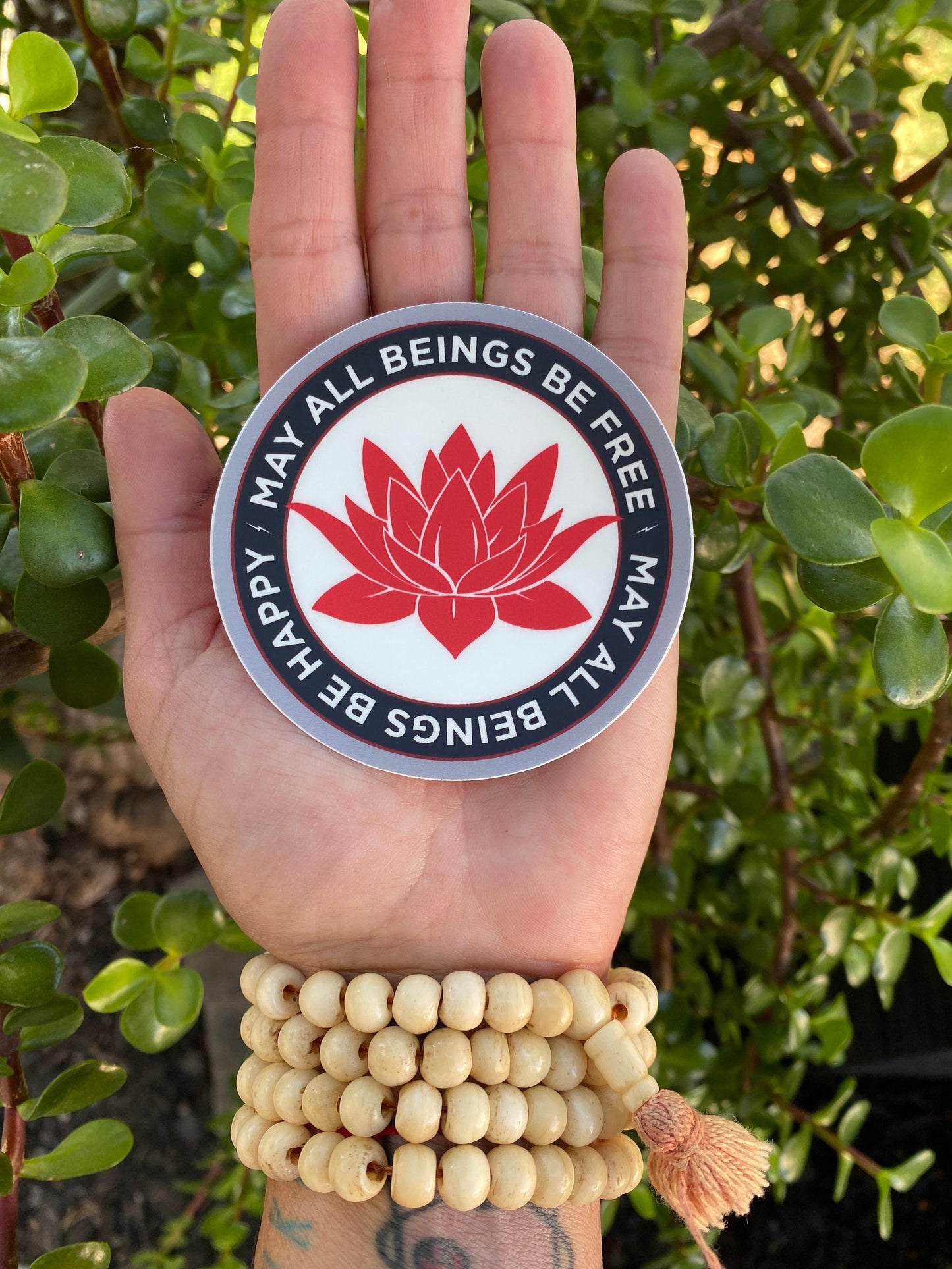 Hand holding a circular sticker with a red lotus flower design and text, may all beings be free may all beings be happy, against a green leafy background. wearing a brown mala, prayer beads.