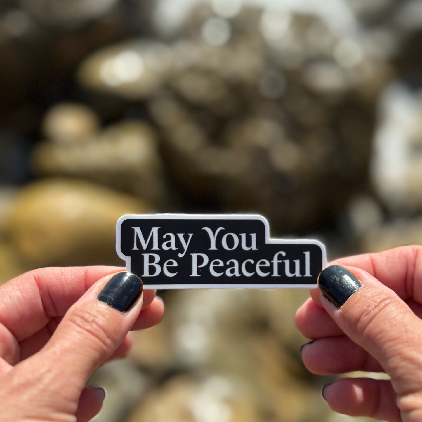 Hand holding a Buddhist sticker with 'May You Be Peaceful' text against a blurred natural background