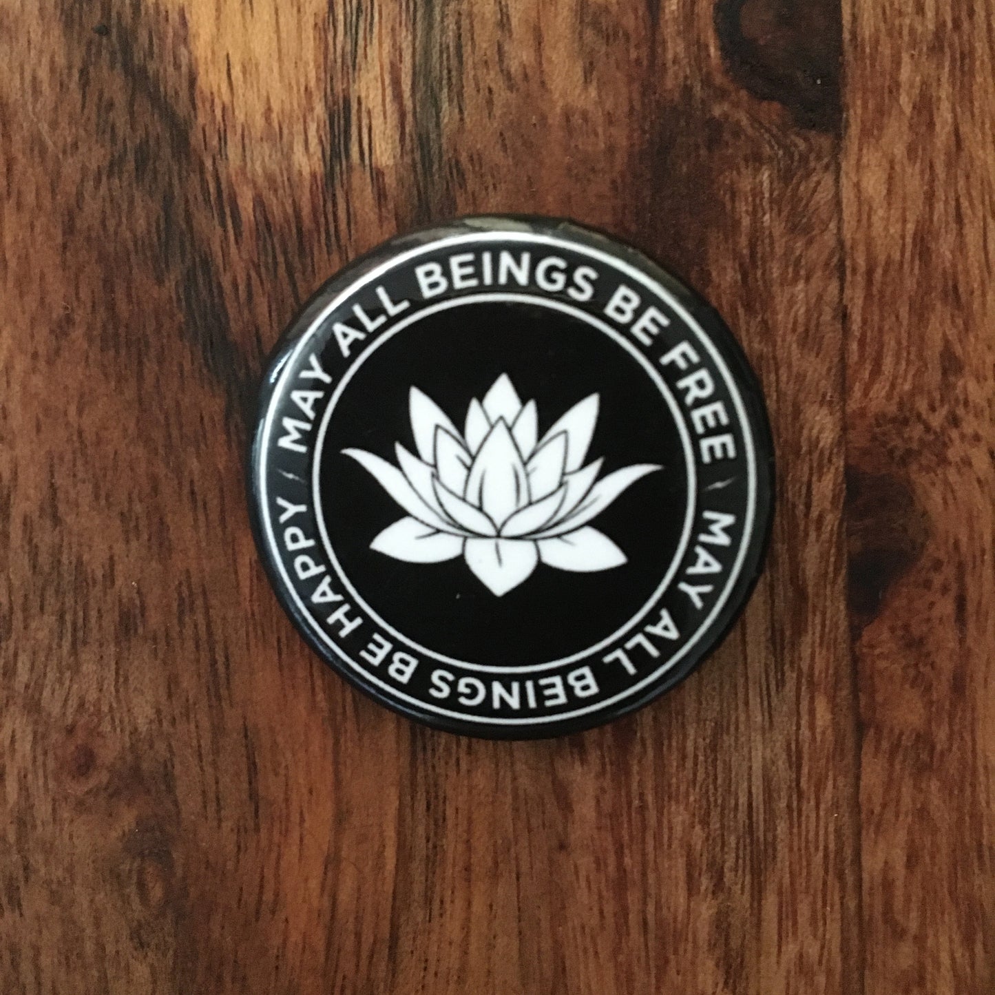 Round 1.25" Buddhist black and white pin back button with a lotus flower and text on a wooden surface.