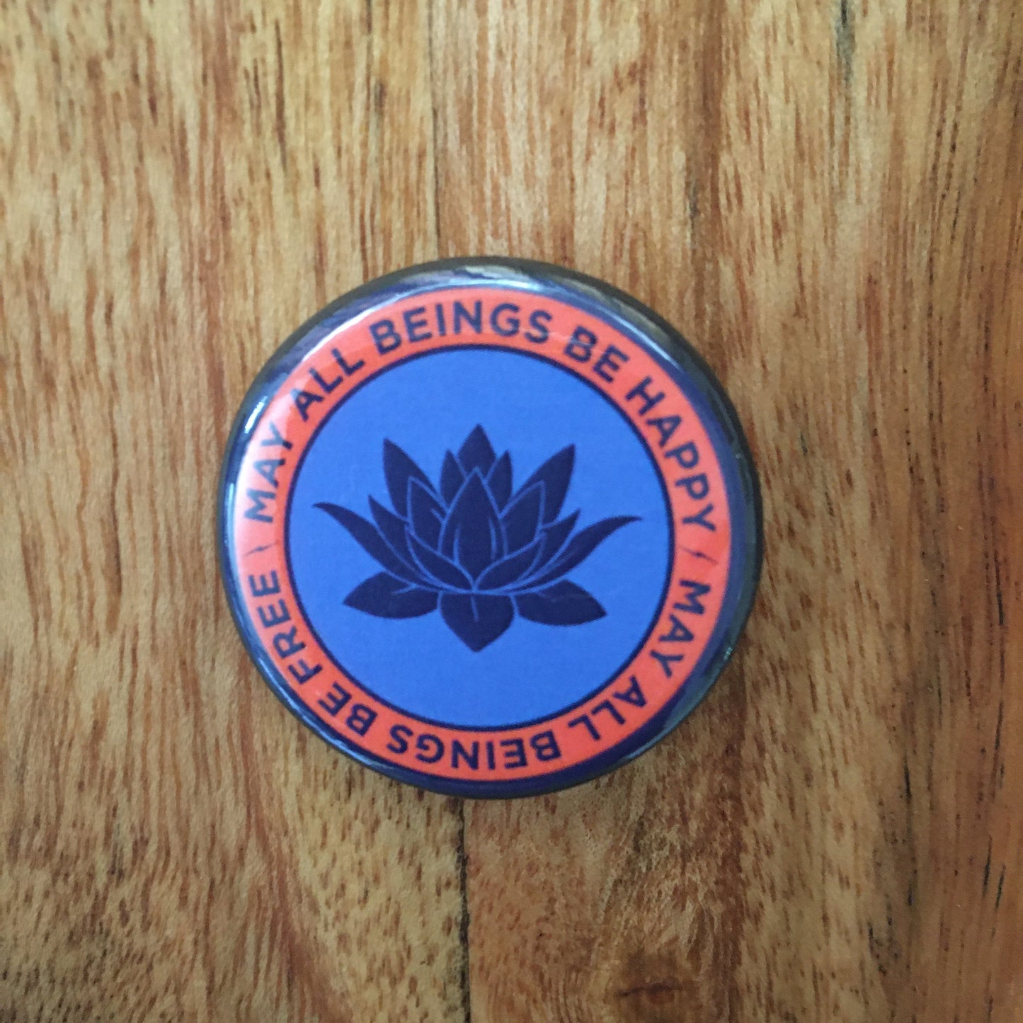 1.25" circular pin back button with a lotus flower design and text, may all beings be free may all beings be happy, text on a wooden surface.
