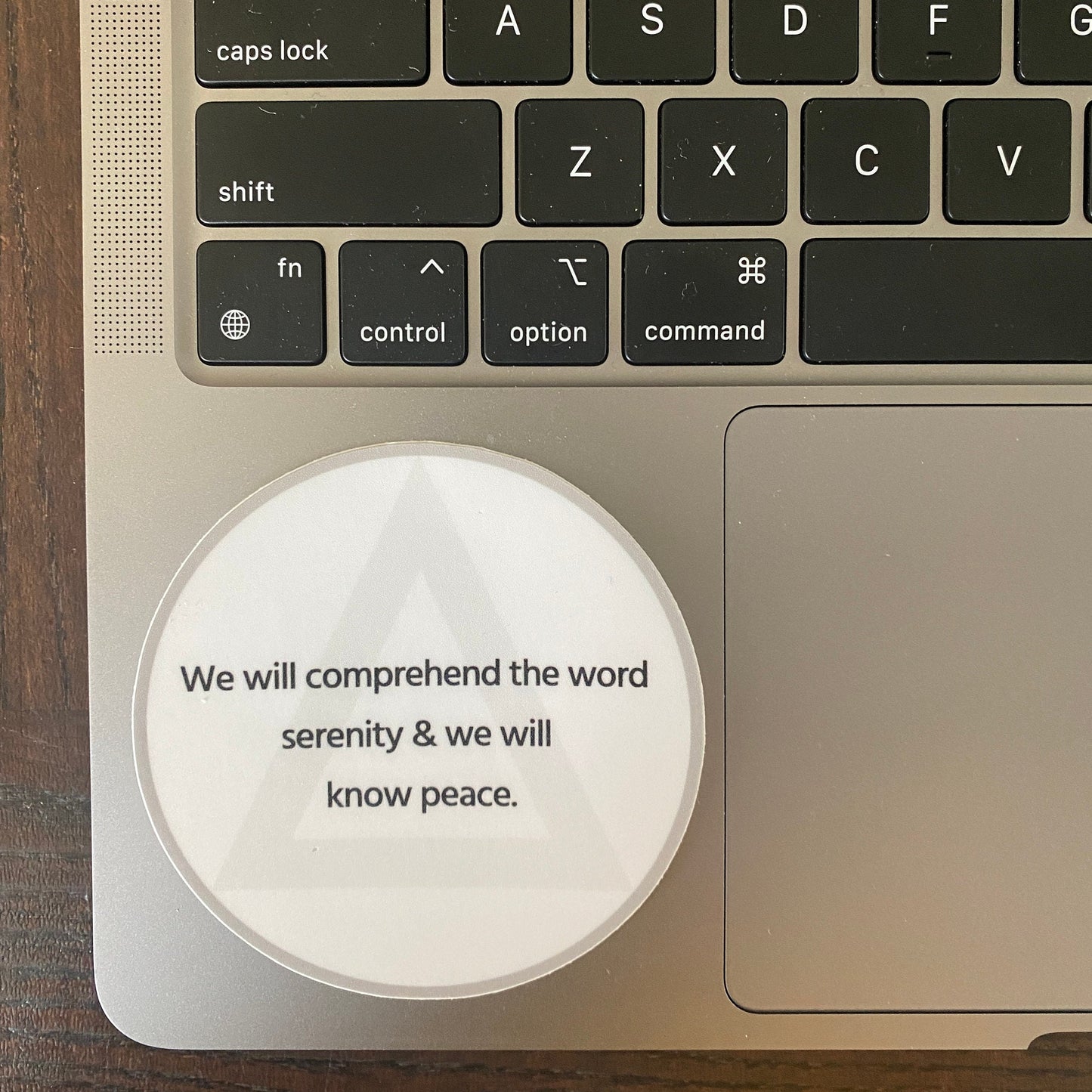 Laptop trackpad with three inch round white sticker with AA circle and triangle design and the quote, we will comprehend the word serenity and we will know peace.