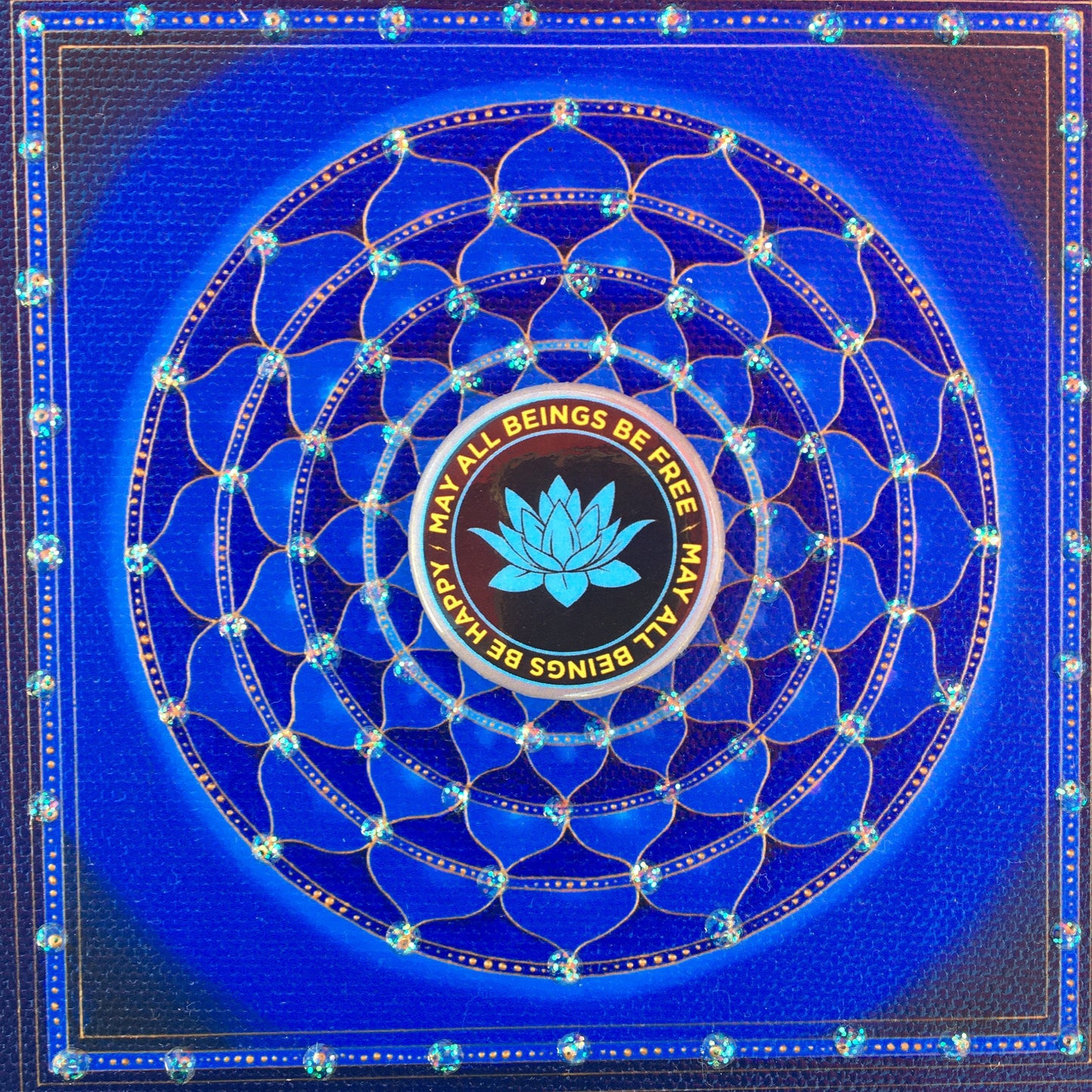 Blue mandala with a 1.25" black and blue lotus button with the Buddhist prayer, may all beings be free may all beings be happy, in the center.