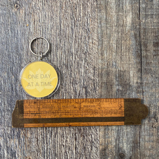 Keychain with motivational quote, 12 step recovery slogan 'one day at a time' on a wooden ruler against a wooden background.
