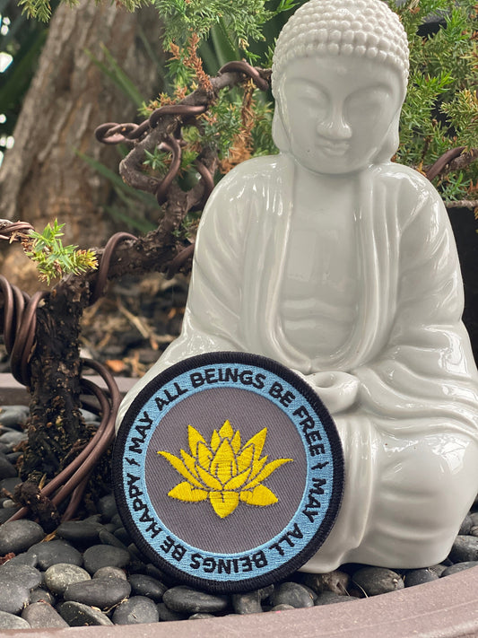 White Buddha statue with 3" round embroidered golden lotus flower design and the text, may all beings be free and may all beings b e happy, a pebble surface.