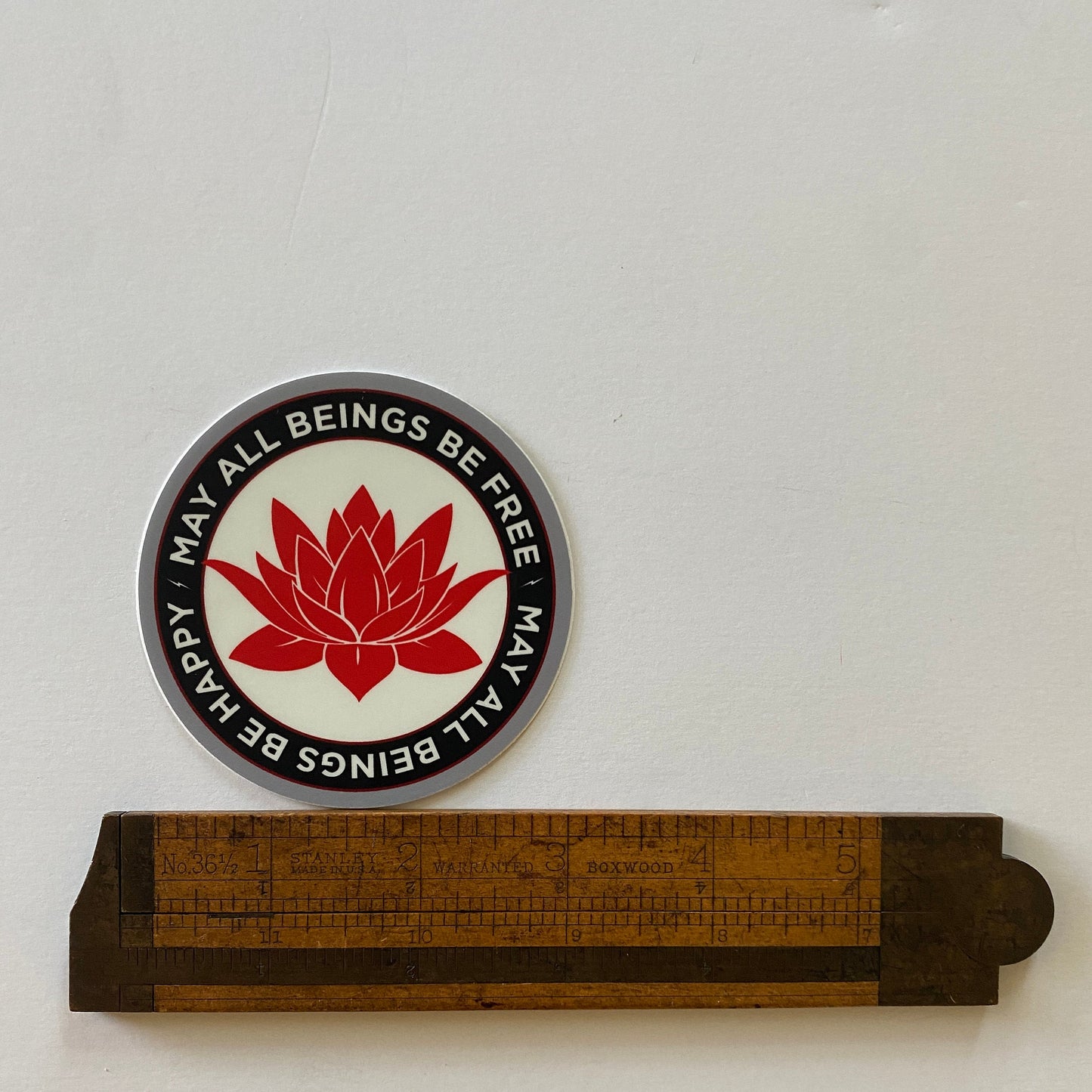 Circular sticker with red lotus flower and text 'May all beings be free' on a white background, with a wooden ruler below.