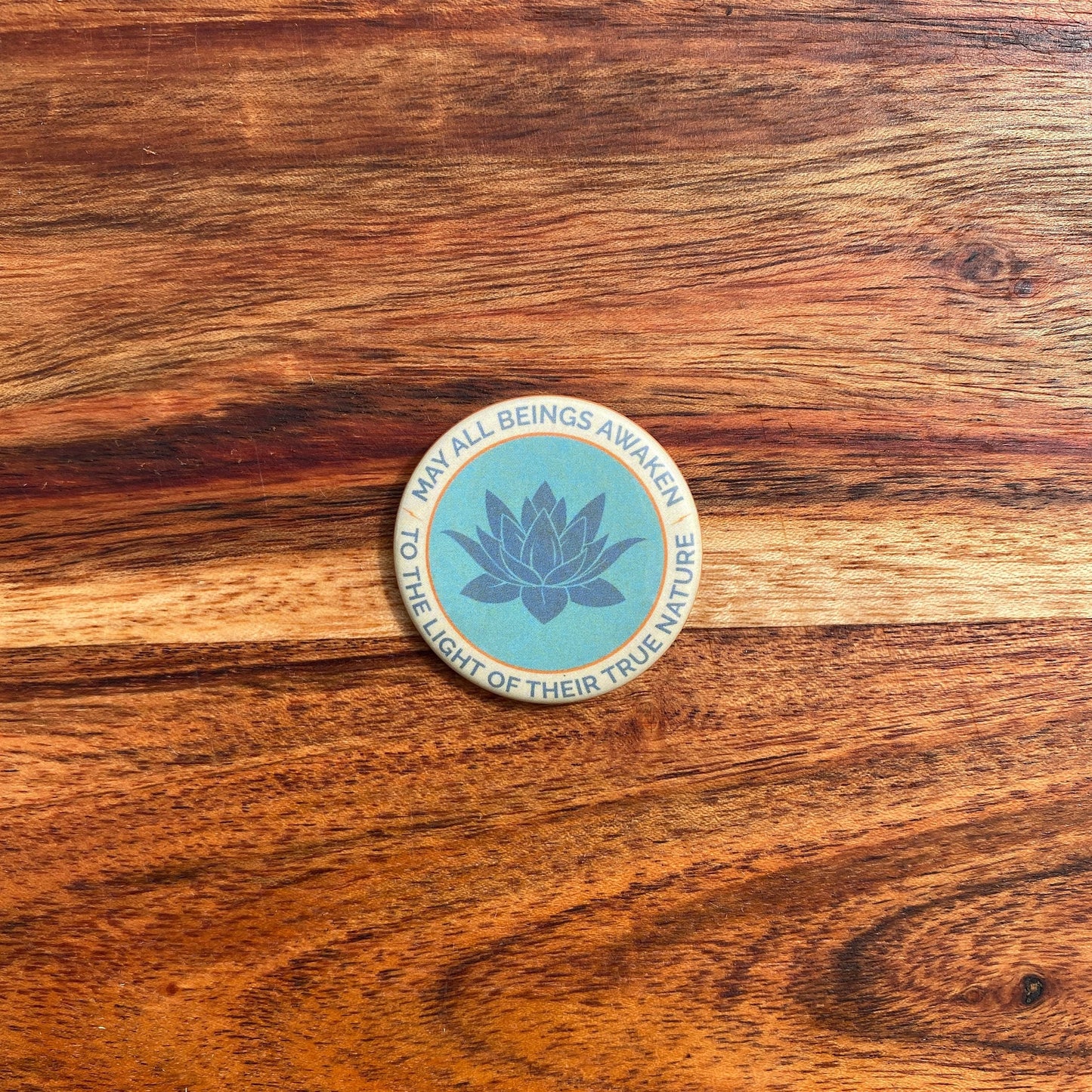 1.5" button with the mantra that reads, may all beings awaken to the light of their true nature, with a lotus design design on a wooden surface