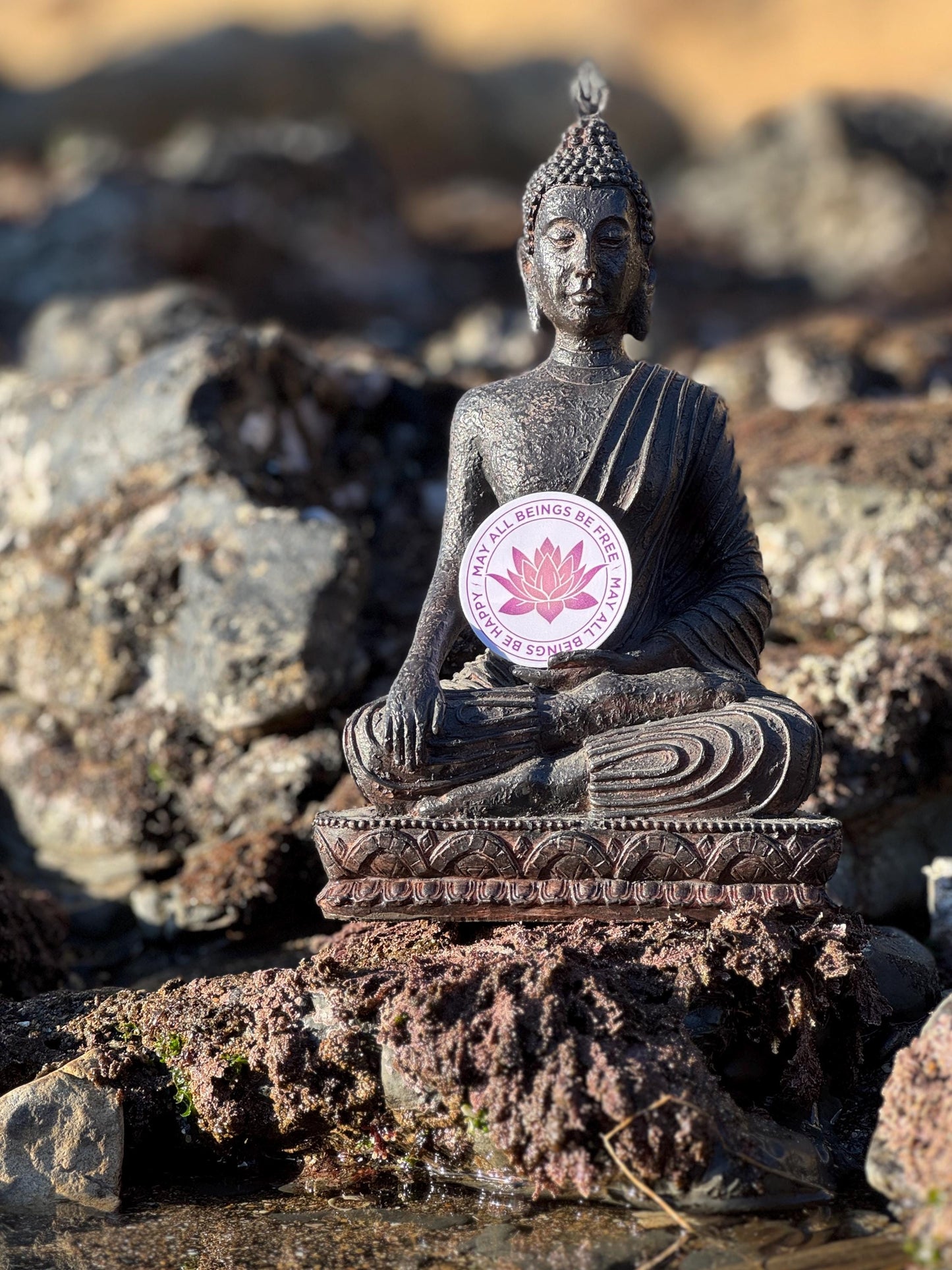 Buddha statue on rocks with a 2" purple sticker with a lotus design and the text, may all beings be free may all beings be happy.