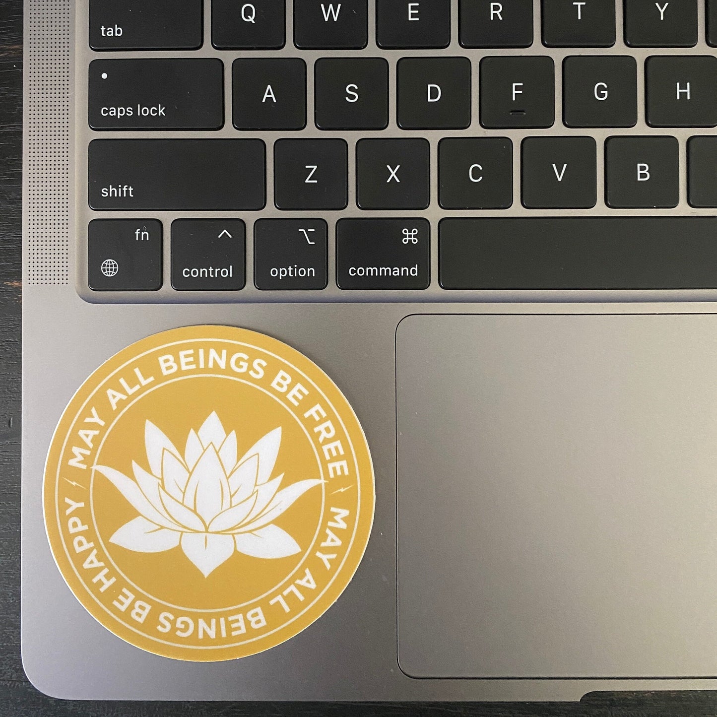 Laptop with a 3" gold and white sticker with a lotus flower design and text, may all beings be free may all beings be happy, on the palm rest area.