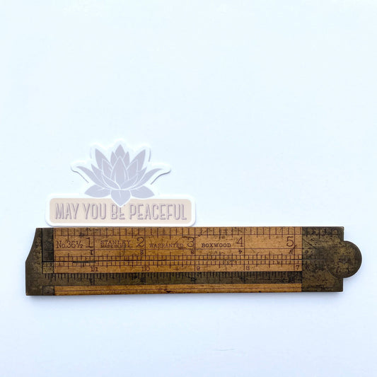 Wooden ruler with 'May You Be Peaceful' message and lotus flower on a white background.