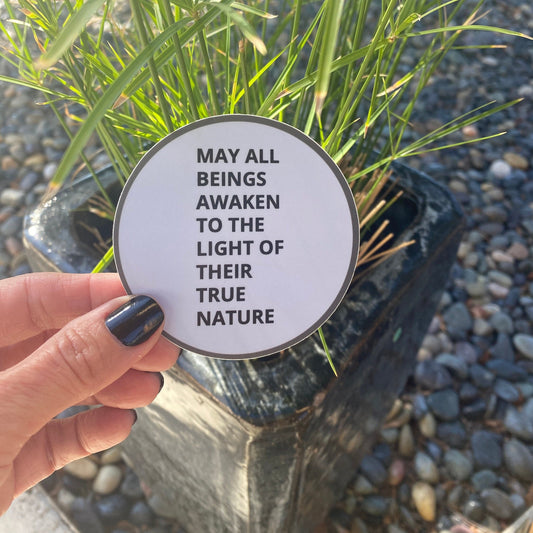 Hand holding a circular sticker with the text, may all beings awaken to the light of their true nature, front of a plant on pebbles