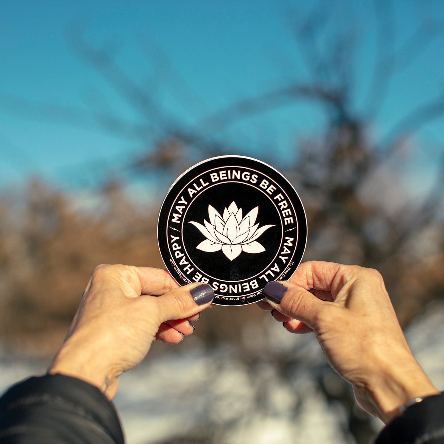 Two hands holding a 4" black and white circular sticker with a lotus flower design and text, MAY ALL BEINGS BE FREE MAY ALL BEINGS BE HAPPY, against a blurred natural background.