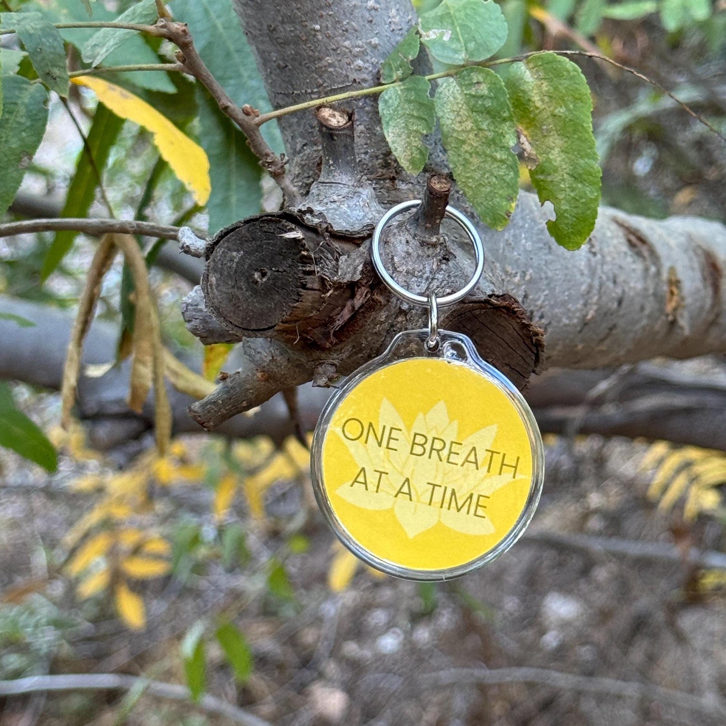 2" yellow lotus design keychain with 'One Breath at a Time' text hanging on a tree branch.