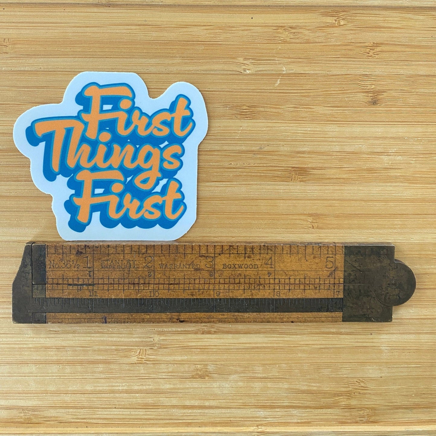 Sticker with 'First Things First' text on a wooden surface with a ruler for scale.