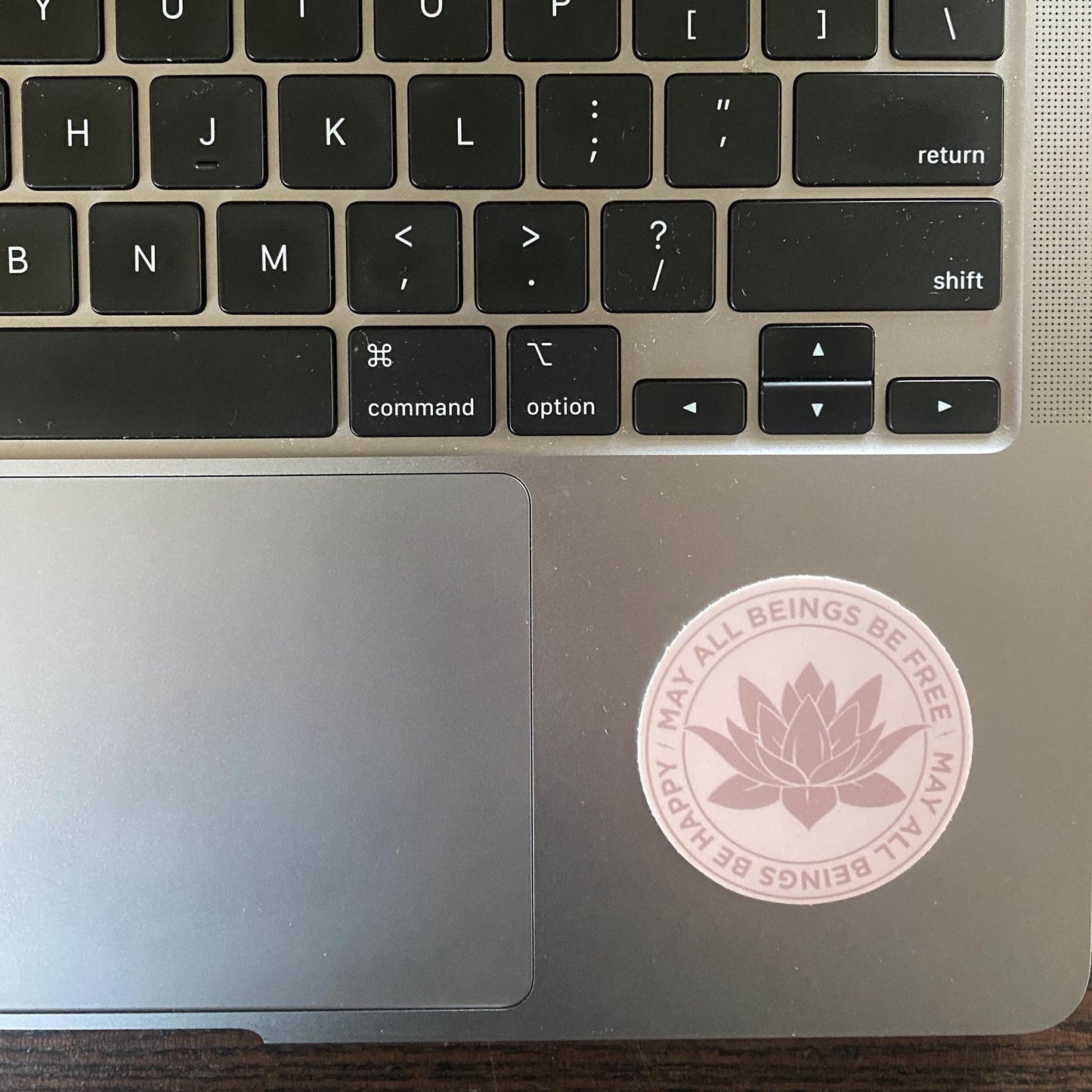 Close-up of a laptop keyboard with a 2" circular pink Buddhist lotus flower sticker that reads, may all beings be free may all beings be happy, on the palm rest area.