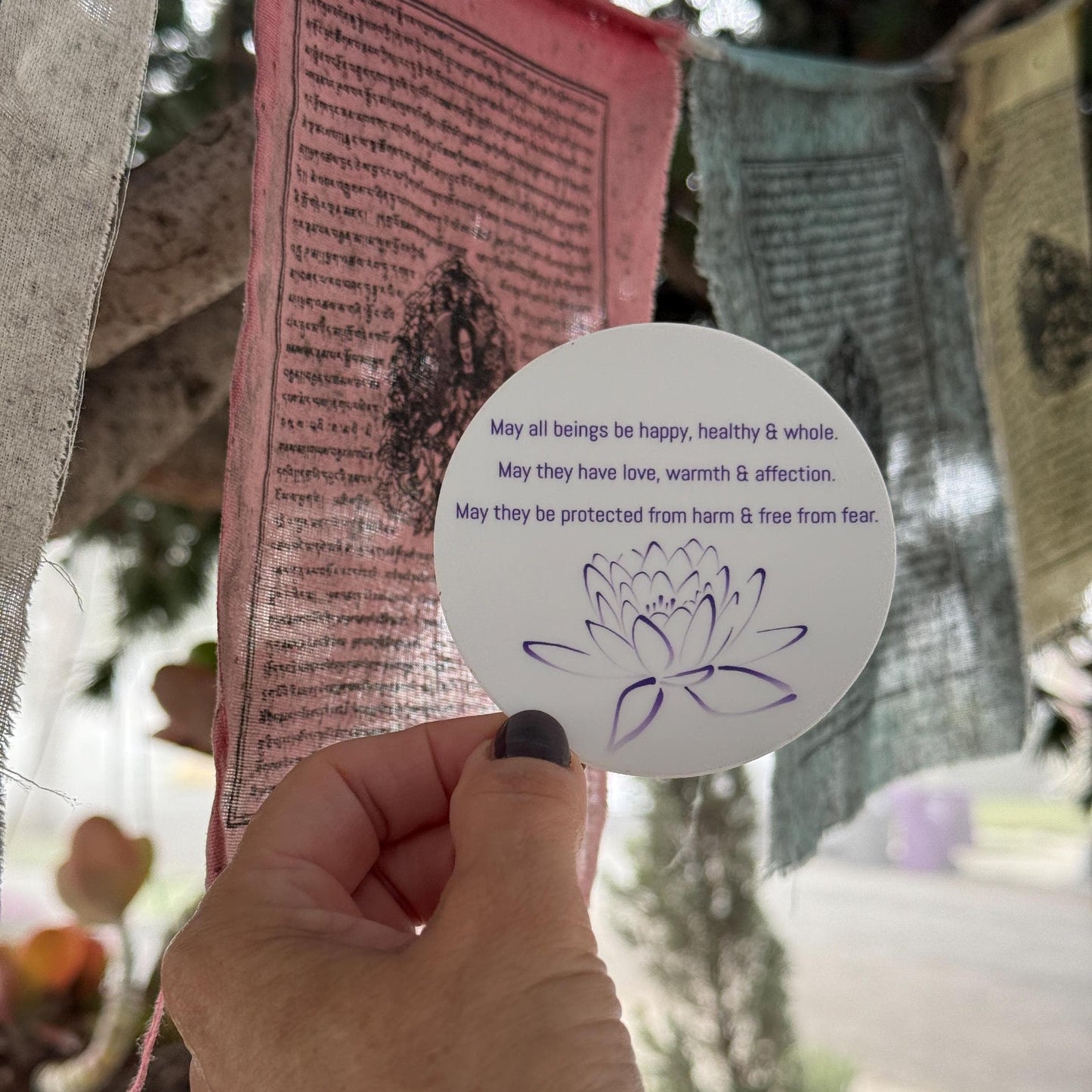 Hand holding a 3" circular sticker with Buddhist prayer and a lotus design in front of prayer flags.