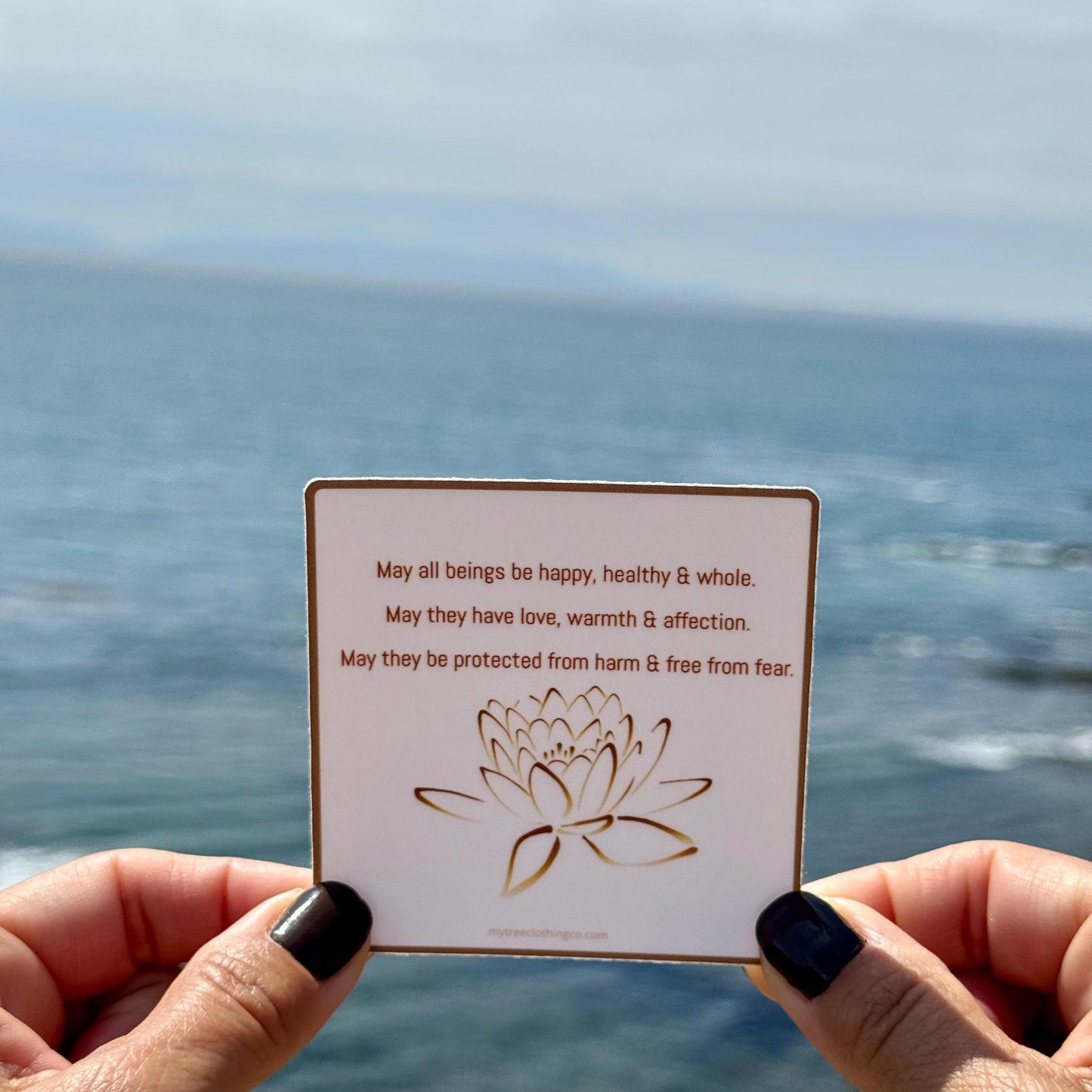 3" square Buddhist sticker with a lotus flower design and text, 'May all beings be happy, healthy and whole. May they have love, warmth and affection. May they be protected from harm and free from fear.' Held by hands against the Pacific Ocean background.