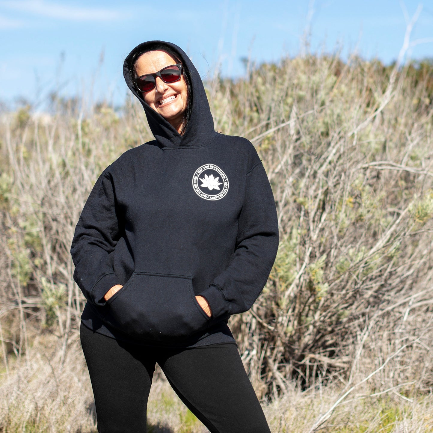 Person wearing a black hoodie with a white lotus and the mettā prayer, 'may you be peaceful may you be happy may you be free.' Standing outdoors in a natural setting.