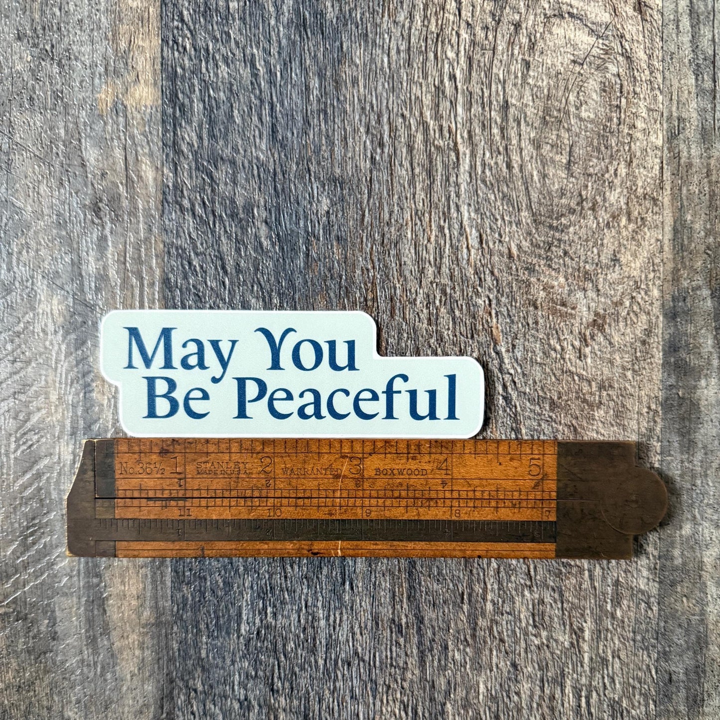 Wooden ruler with light blue and dark blue 'May You Be Peaceful' sticker on a wooden background.