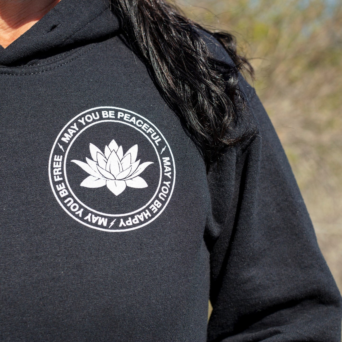 Close up of front of black hoodie with a white lotus and the mettā prayer, 'may you be peaceful may you be happy may you be free.'
