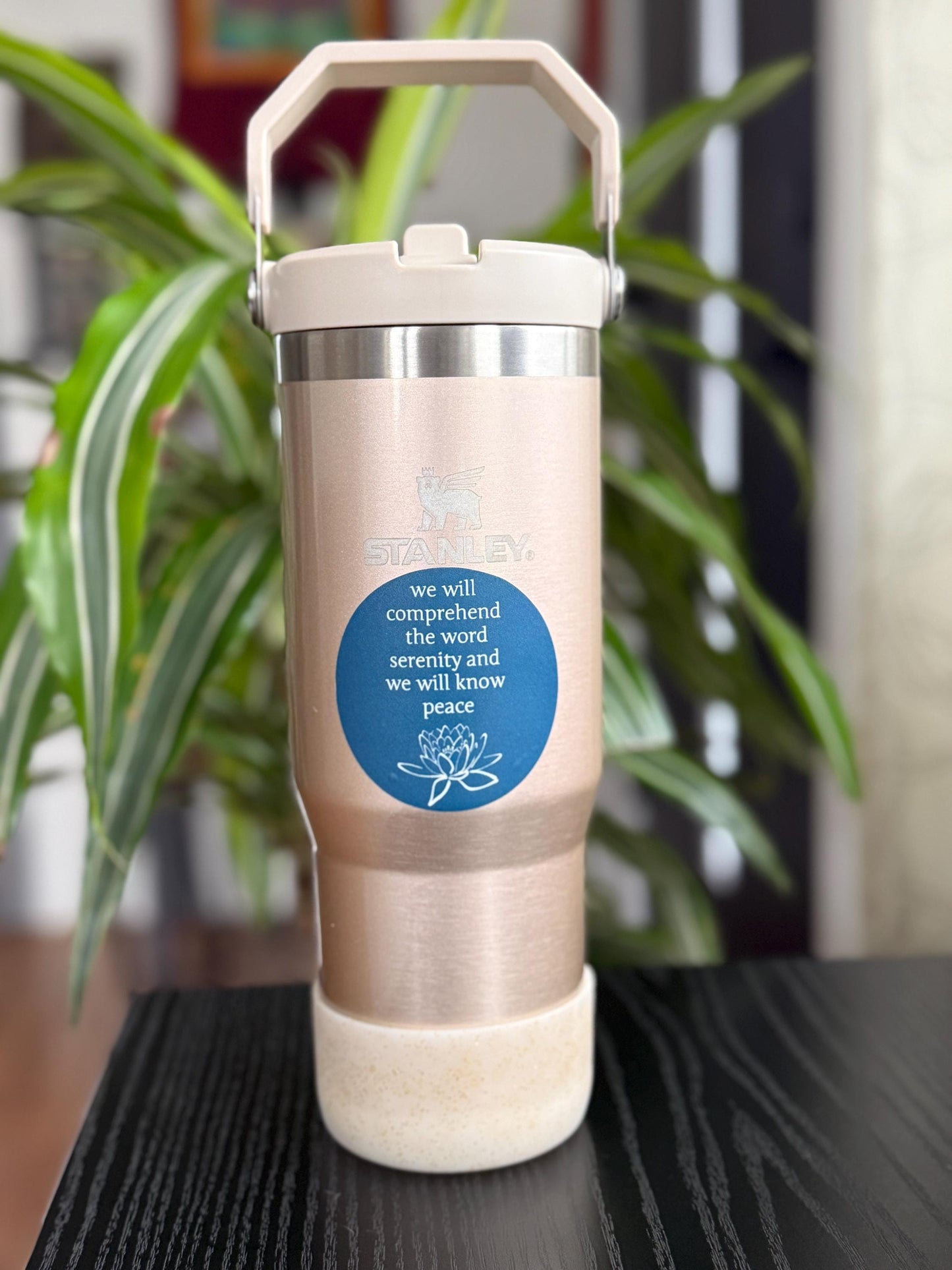 Rose gold tumbler with a blue 2.5" blue sticker with a quote from the big book of AA, "we will comprehend the word serenity and we will know peace" and a lotus flower with a plant in the background