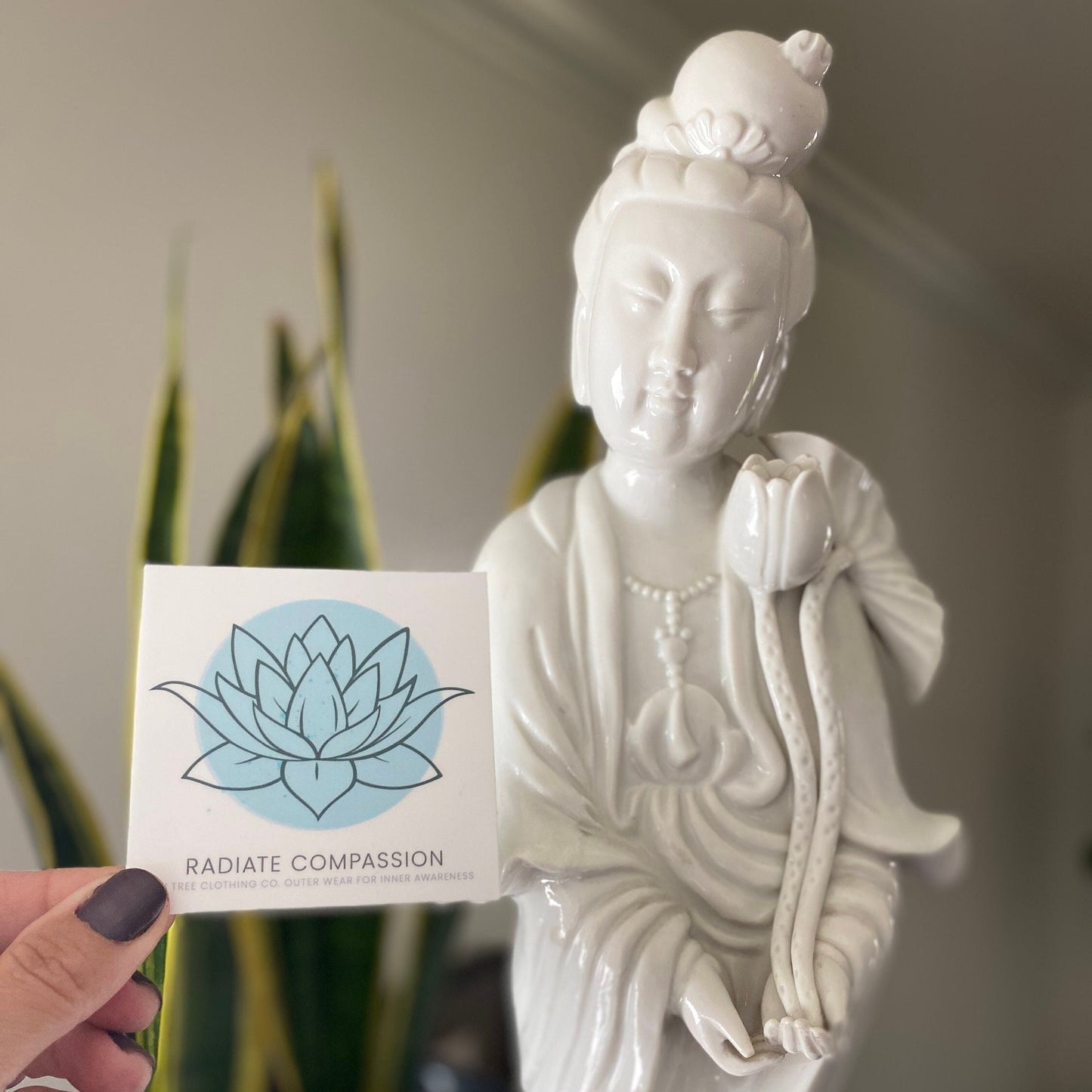 White statue of a Bodhisattva seated figure holding a 3" square sticker with a blue lotus design and text, radiate compassion.