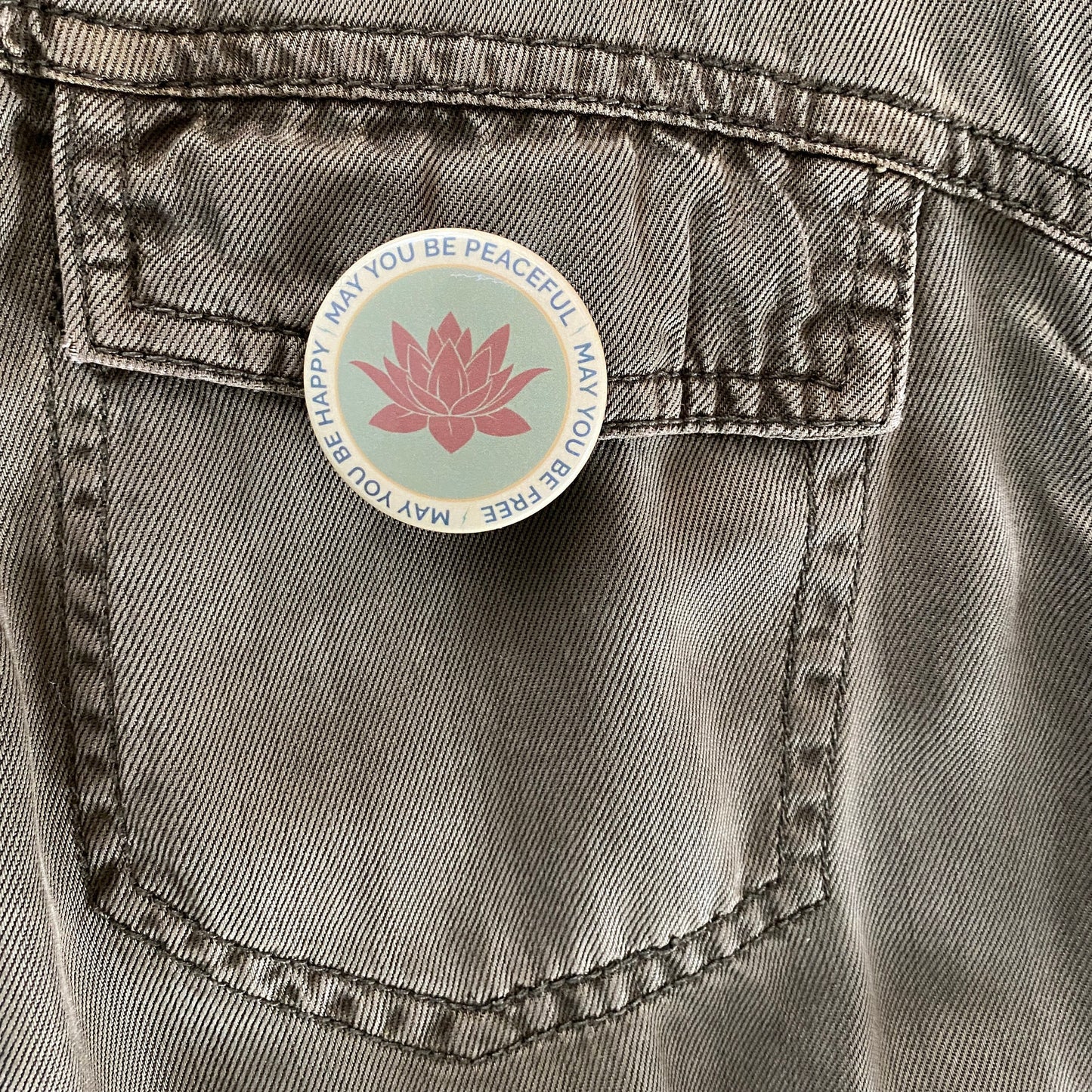 Button with a pink lotus flower design and text, may you be peaceful may you be free may you be happy, on a denim pocket.