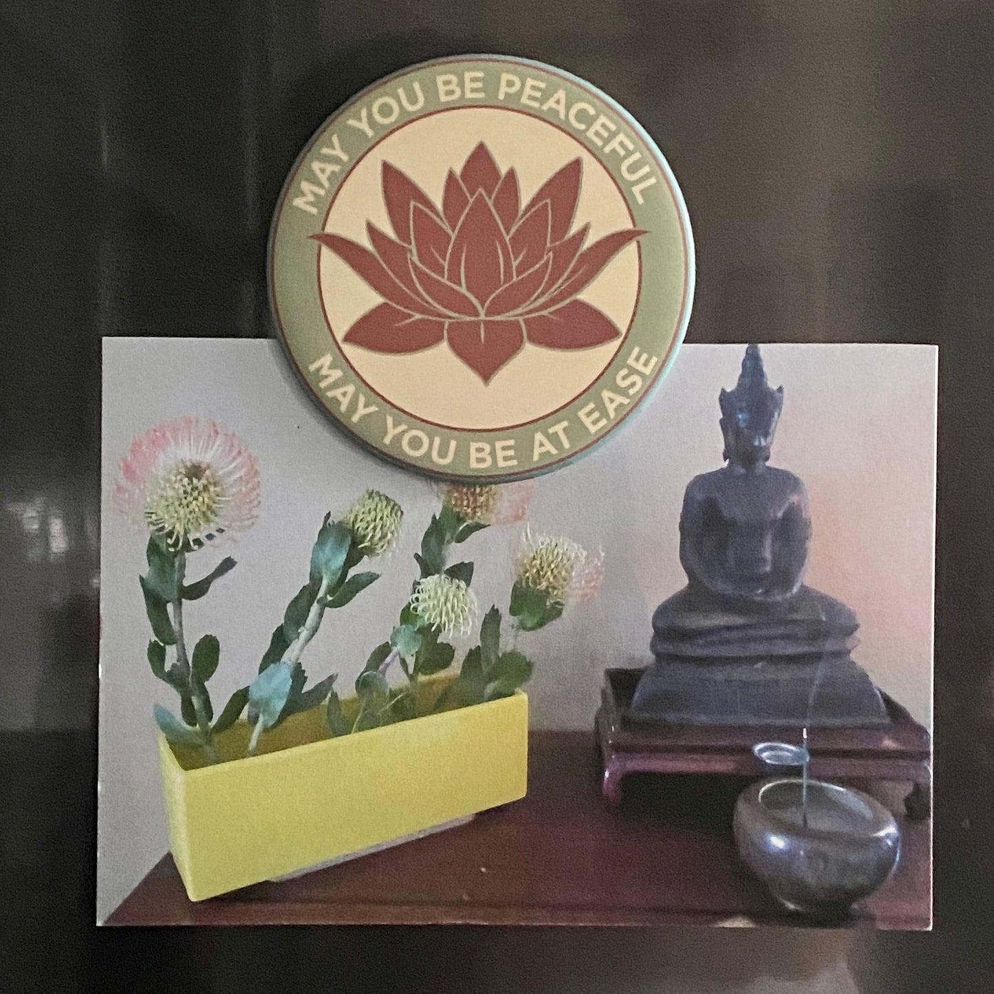 3.5" lotus flower magnet with the metta prayer, 'may you be peaceful may you be at ease' on a refrigerator holding a photo of a Buddha statue, incense, and Ikebana.