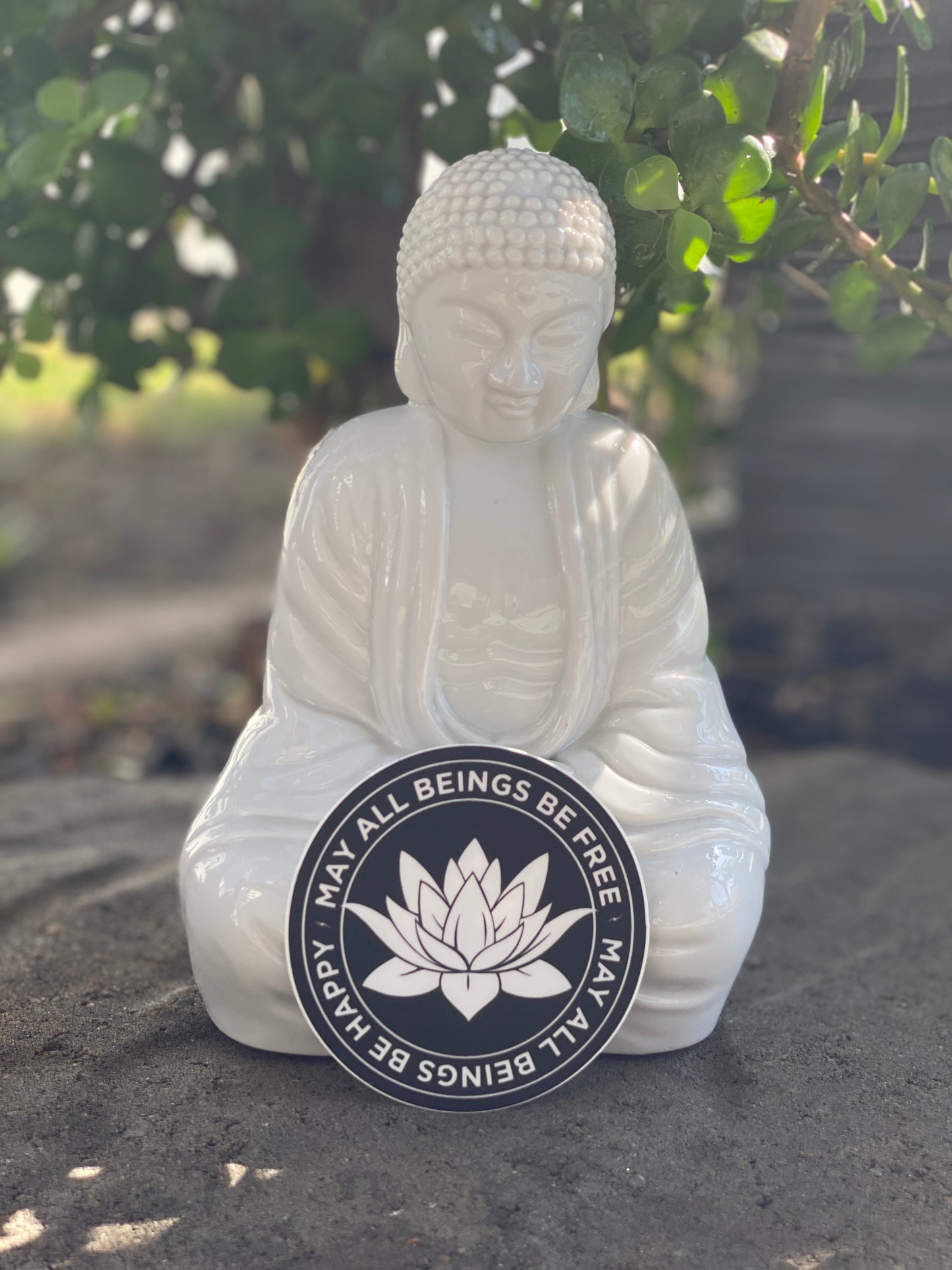 White Buddha statue with a circular emblem featuring a lotus flower and text, may all beings be happy-may all beings be free, on a natural background.