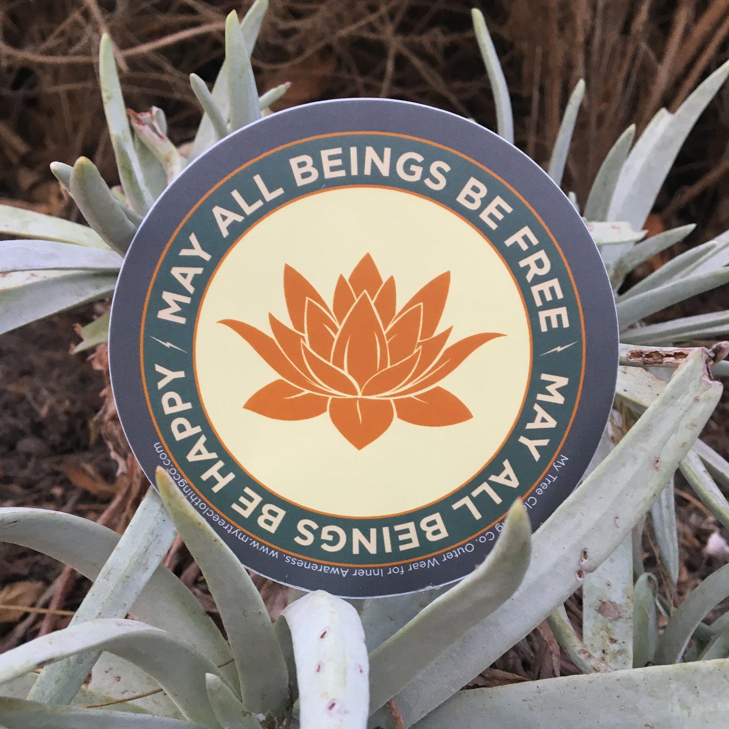 4" circular sticker with a saffron colored lotus flower design and Mettā prayer, held in front of wild sage in the desert of Southern California.