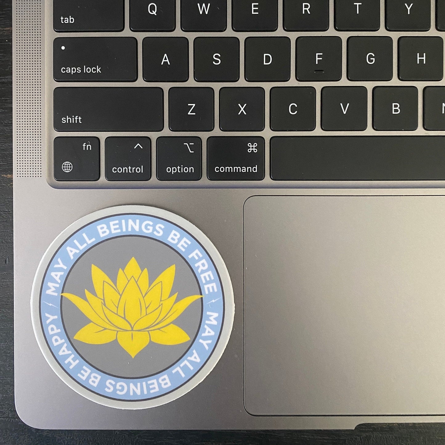Laptop with a 3" circular sticker with a lotus flower design and text, may all beings be free may all beings be happy text on the palm rest area.