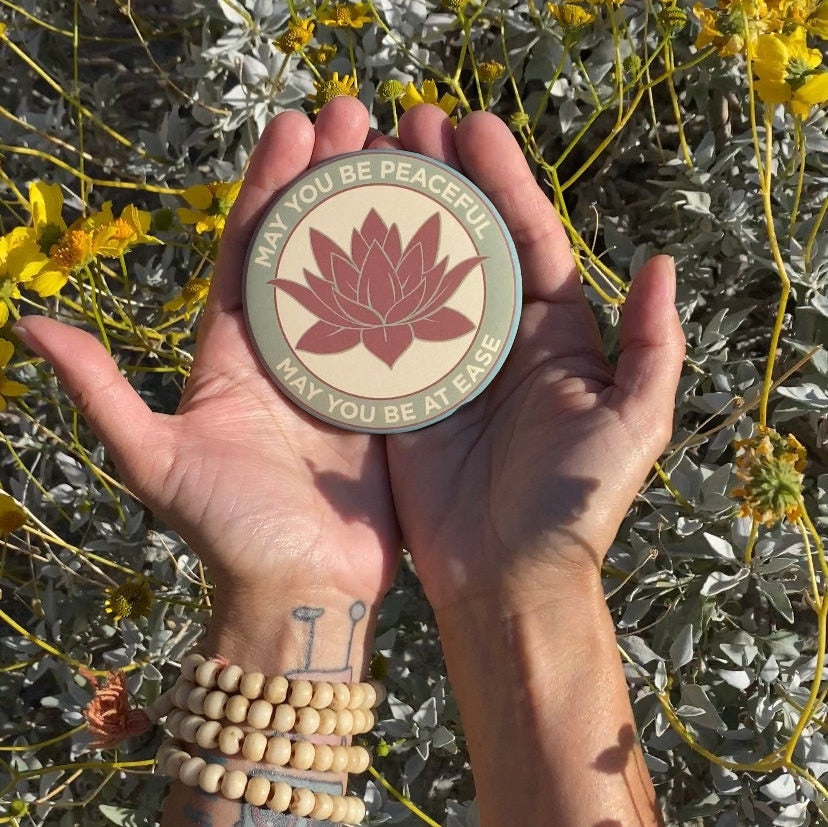 Hand holding a 3.5" lotus flower magnet with the metta prayer, 'may you be peaceful may you be at ease' Surrounded by desert plants.
