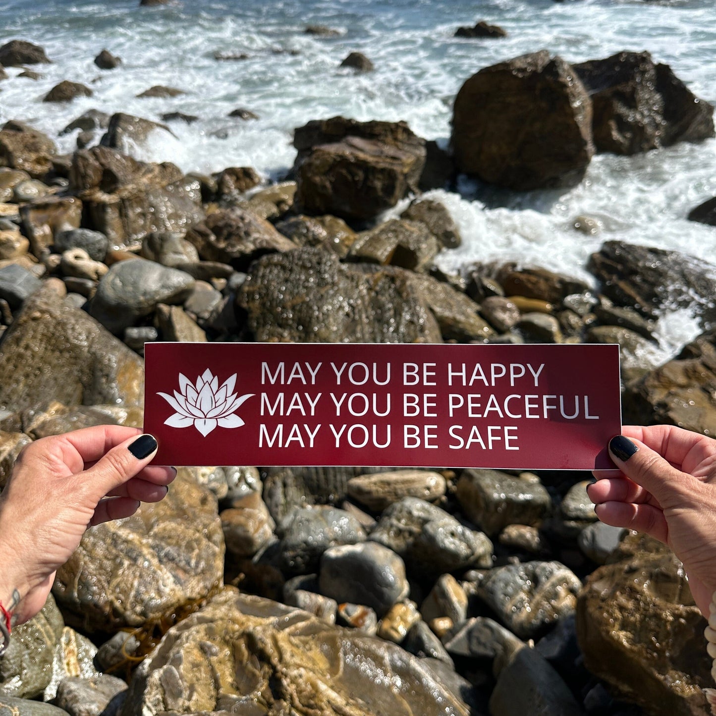 Person holding a red and white bumper sticker with a lotus and the prayer, may you be happy may you be peaceful may you be safe.