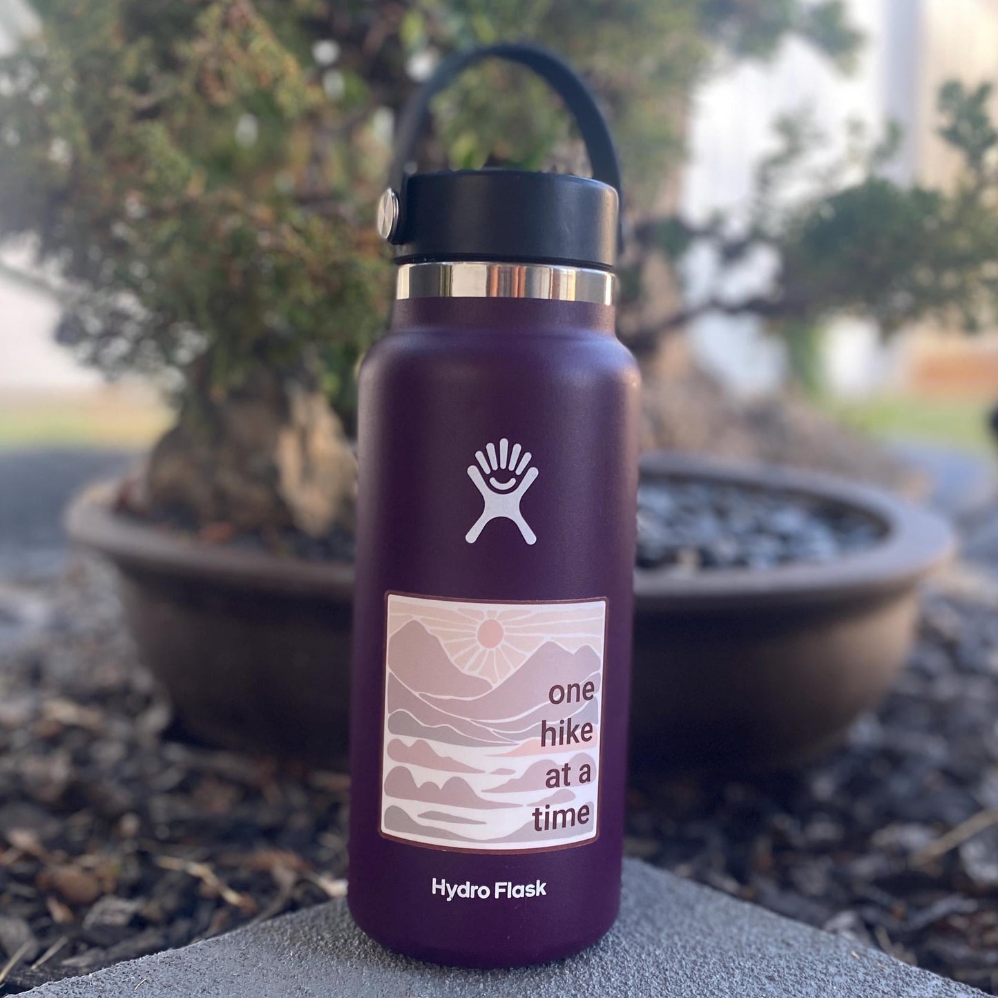 Purple Hydro Flask water bottle with a 3" square sticker with 'one hike at a time' in neutral colors with mountains and a sunset.