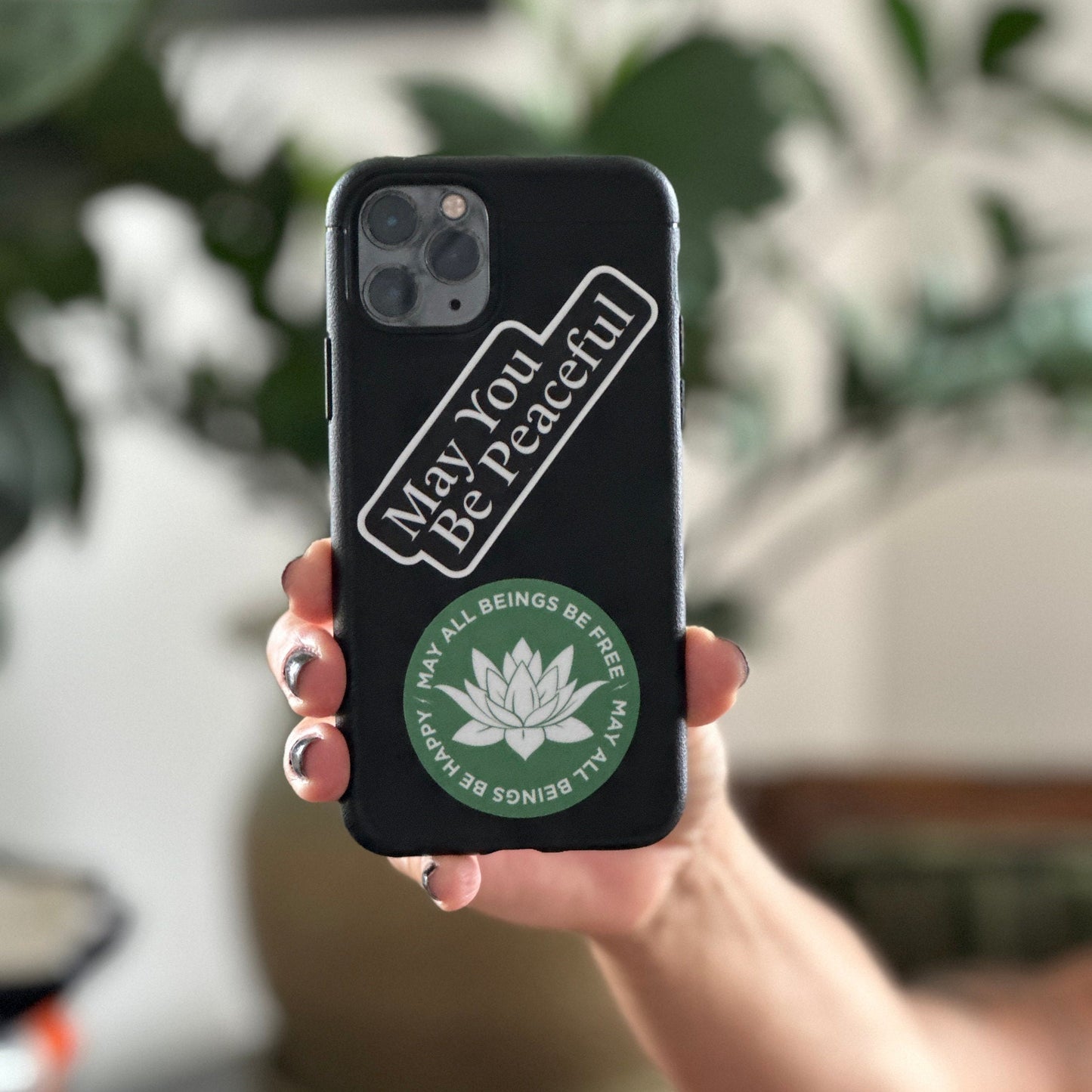 Person holding a phone with a sticker of the mantra 'May You Be Peaceful' on their phone case against a blurred indoor background.
