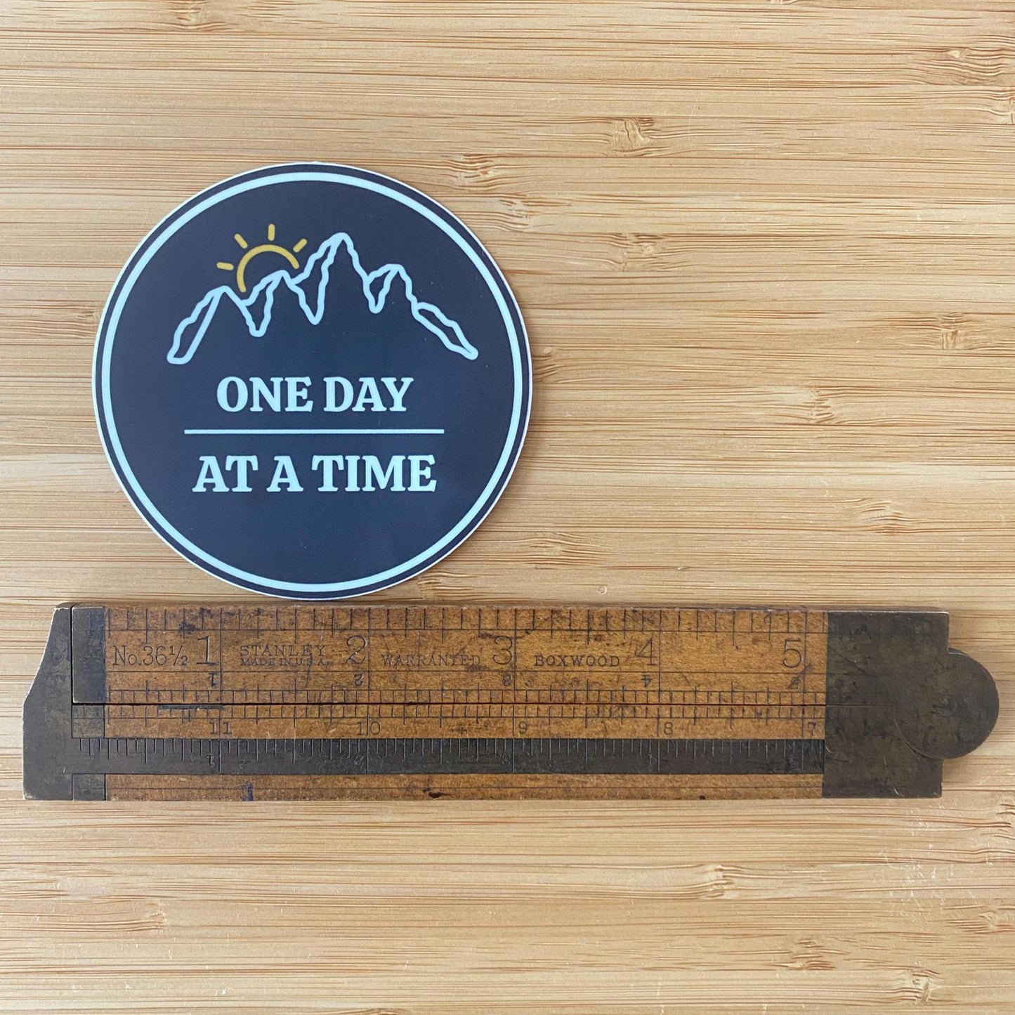 "3 round sticker with outline of mountains and a sunset with 'One Day at a Time' text on a wooden surface with a ruler for scale.