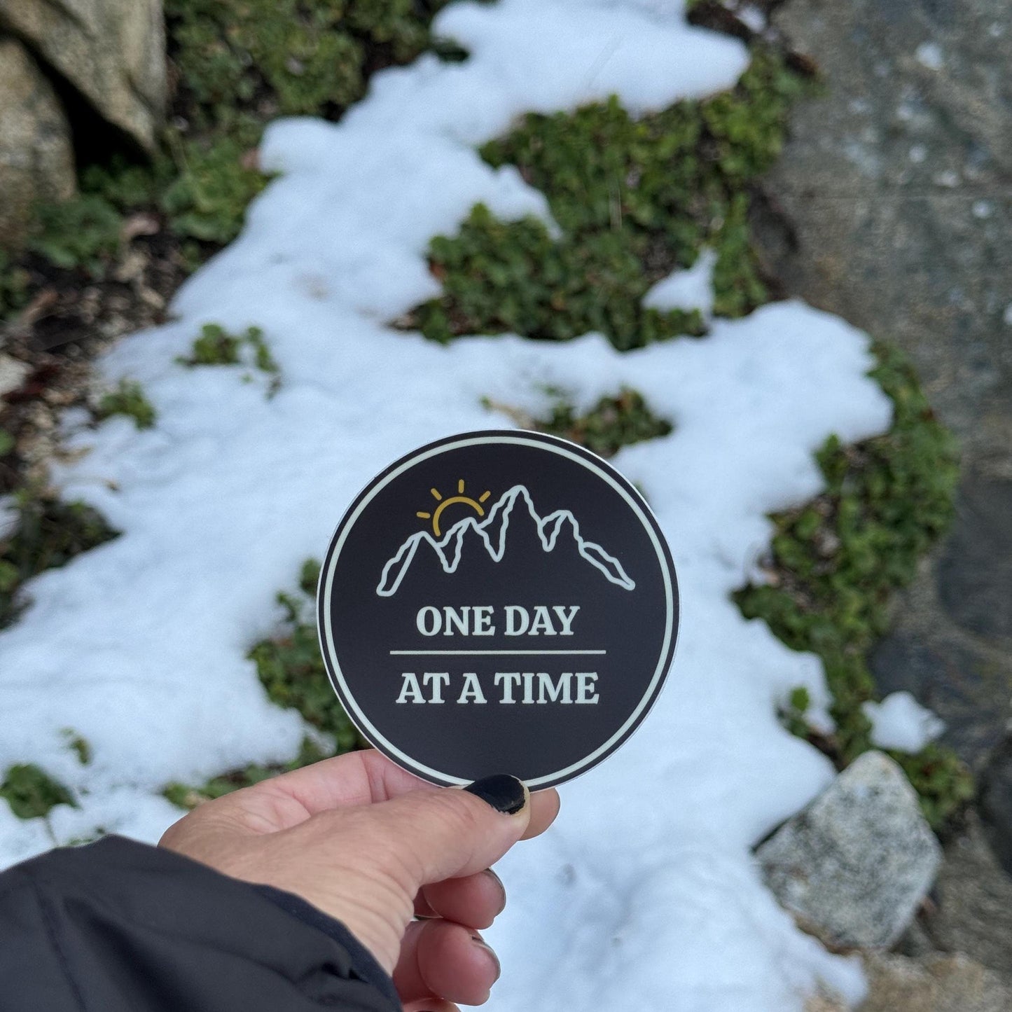 Hand holding a "3 round sticker with outline of mountains and a sunset with 'One Day at a Time' text against a snowy ground background.