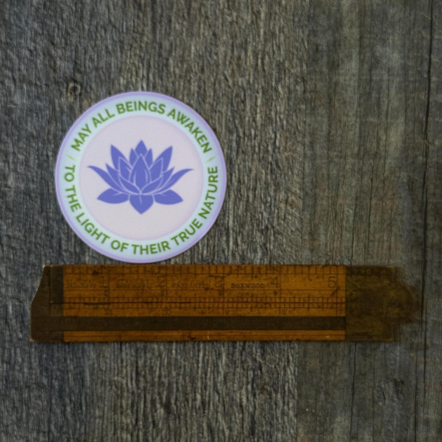 3" round sticker with a purple lotus flower design and the text, may all beings awaken to the light of their true nature. Being measured by an antique wooden ruler.