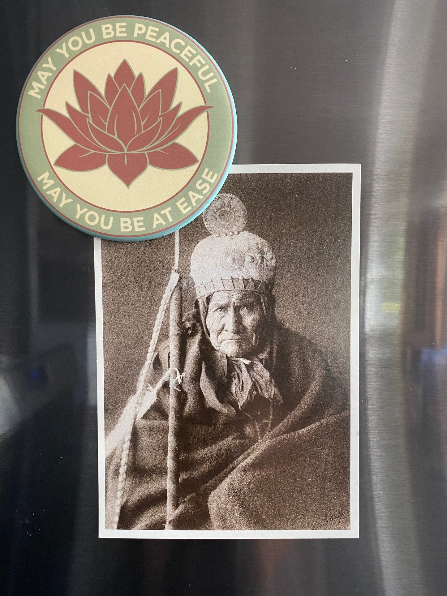 3.5" lotus flower magnet with the metta prayer, 'may you be peaceful may you be at ease' it is holding a photo of Geronimo on a refrigerator.