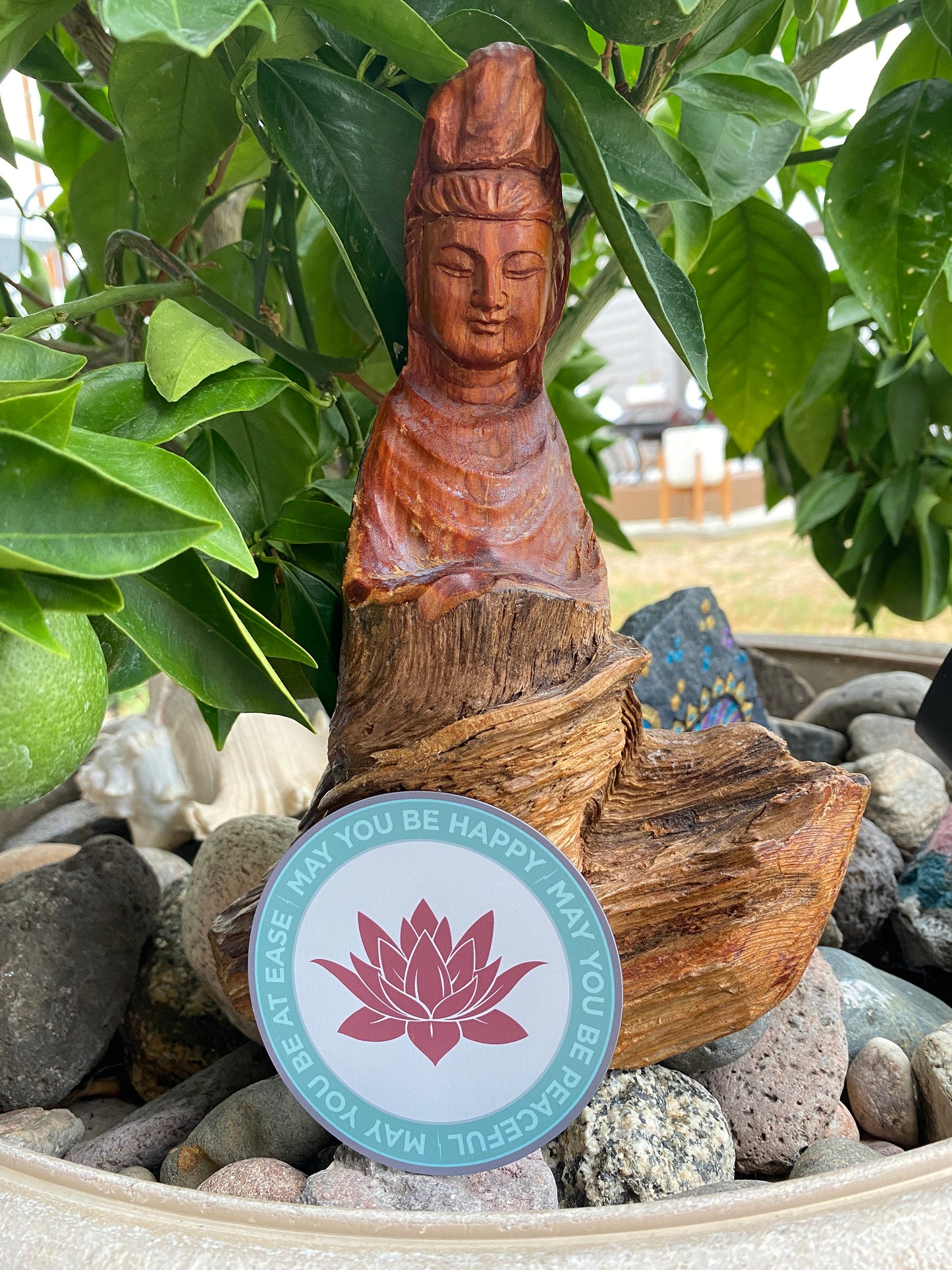 Wooden Bodhisattva statue with a lotus flower sticker design that reads, "may you be happy may you be peaceful may you be at ease" rocks with greenery in the background.