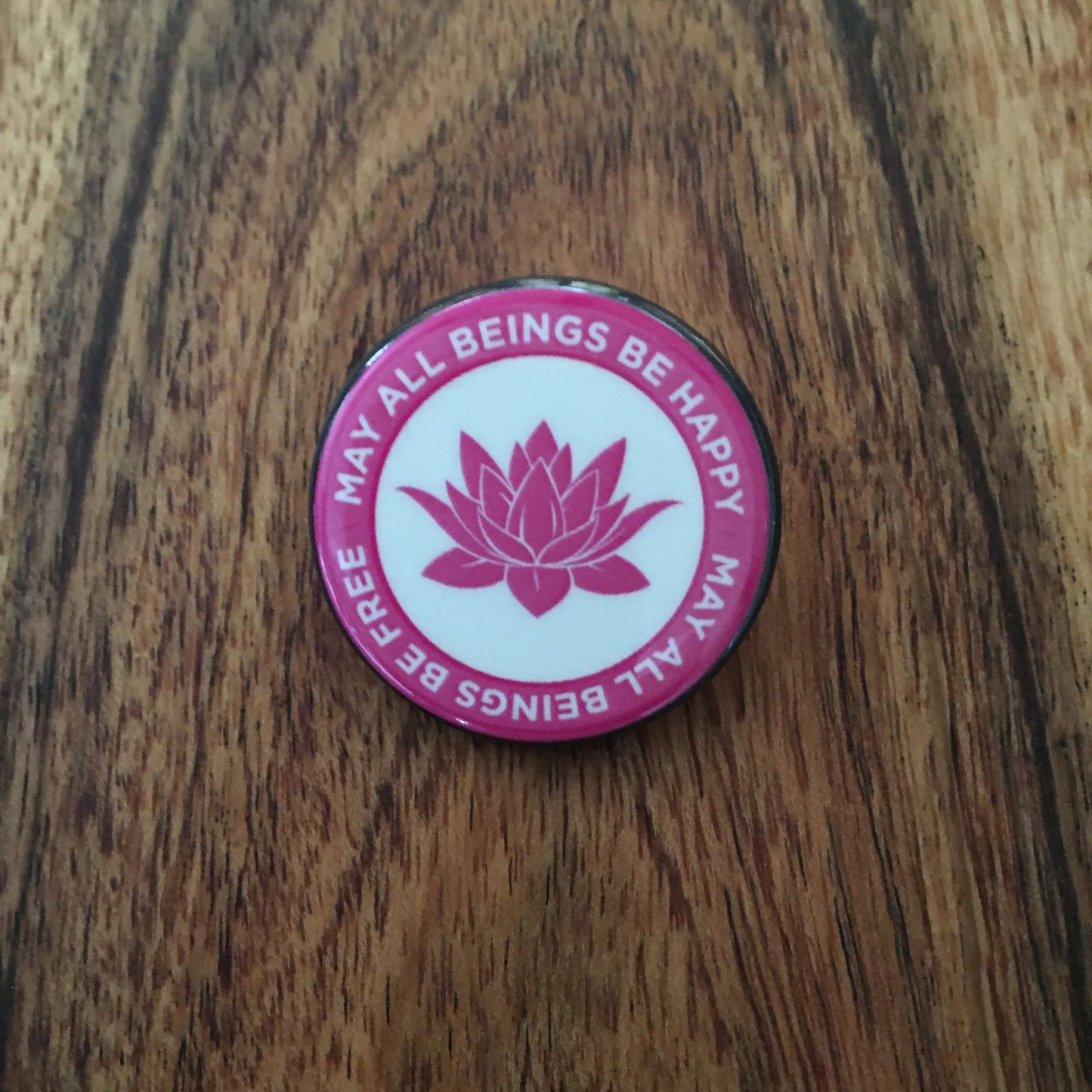 Pink and white circular button with lotus flower and text, Pink button with lotus flower design, may all beings be happy may all beings be free on a wooden surface